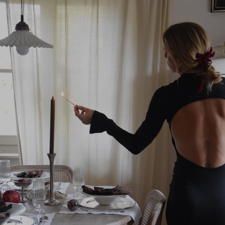Kobieta w czarnej sukience z odkrytymi plecami zapala świecę na elegancko nakrytym stole w jadalni