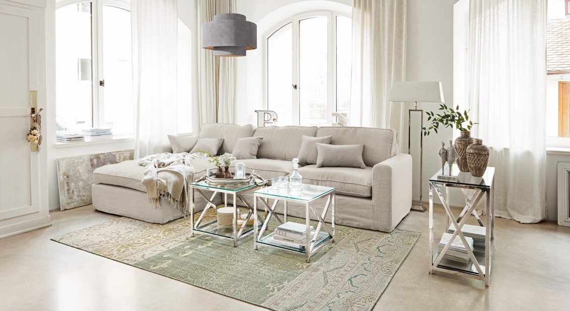Beżowa sofa w lnianym wyglądzie z poduszkami i metalowymi stolikami w jasnym salonie