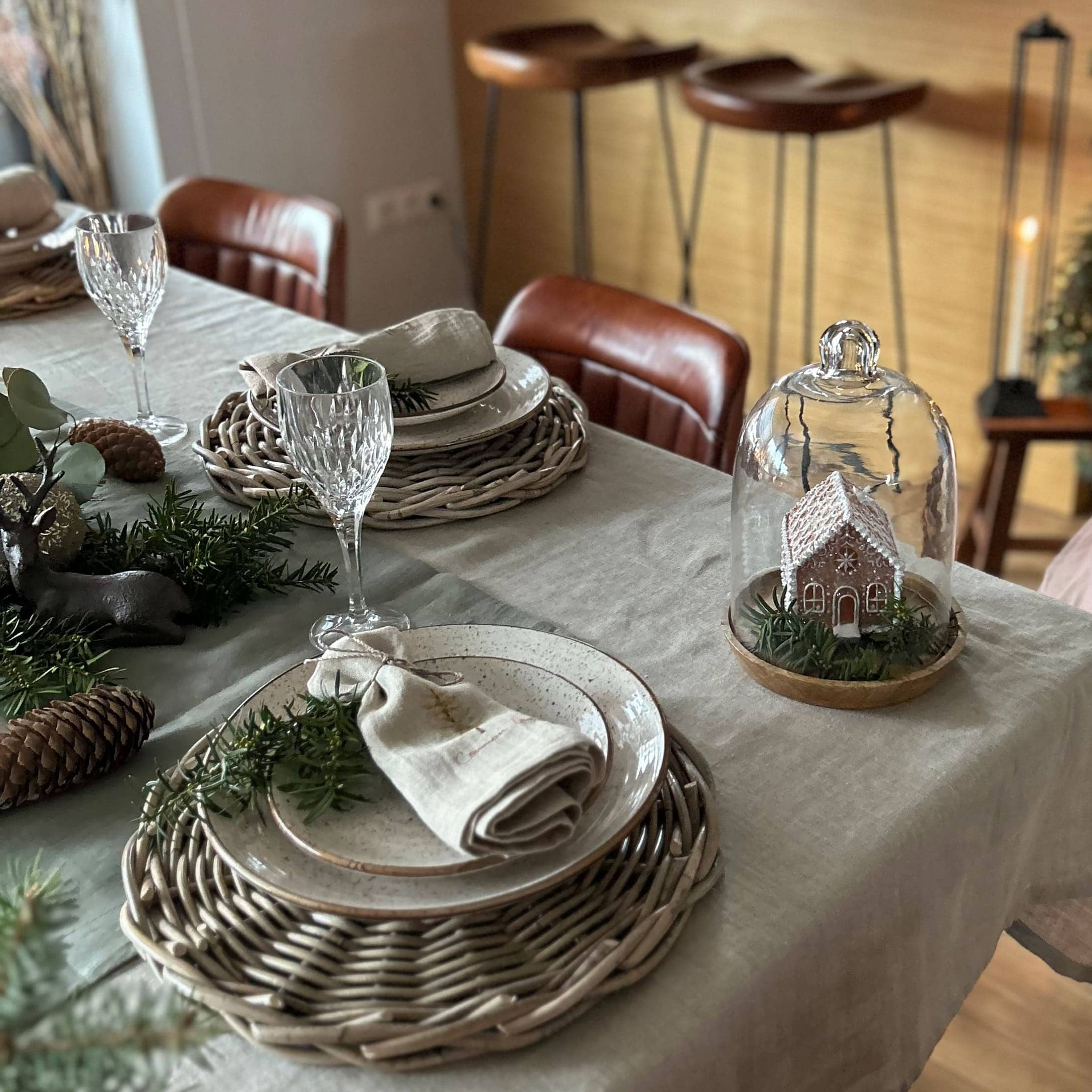 Elegancko nakryty stół z wiklinowymi podkładkami, ceramicznymi talerzami i dekoracją świąteczną z domkiem pod szklaną kopułą