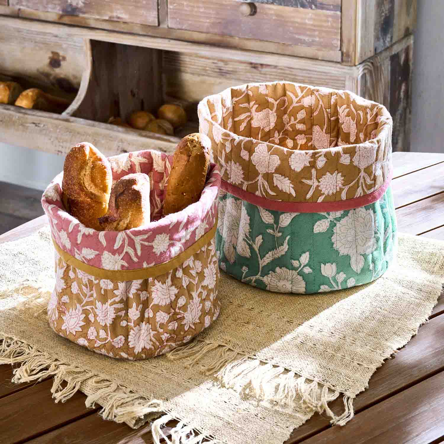 Dwa materiałowe kosze na chleb z kwiatowym wzorem w kolorach różowym, zielonym i brązowym na drewnianym stole