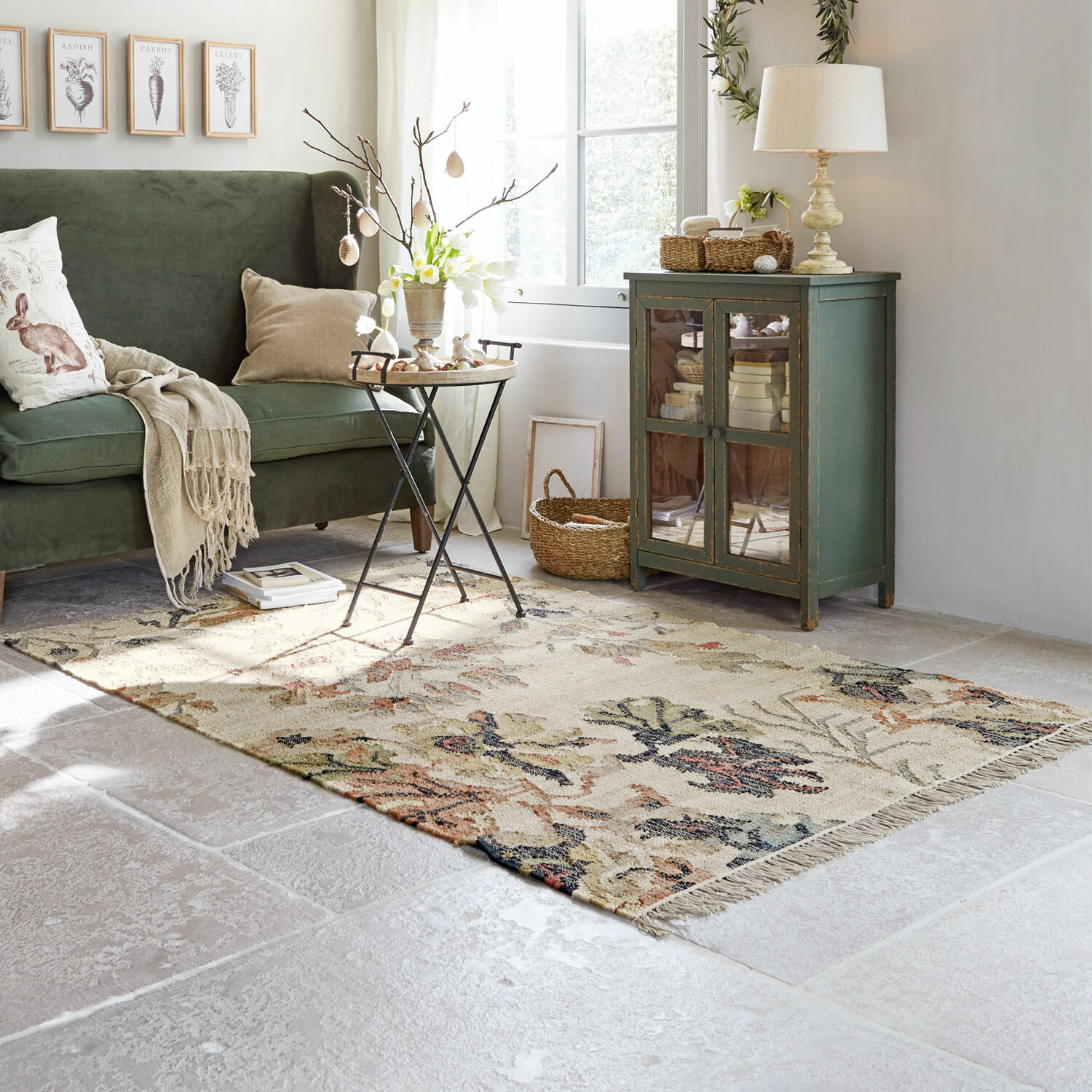 Przytulny salon z zieloną sofą, drewnianą szafką i dywanem w kwiatowy wzór na jasnej podłodze