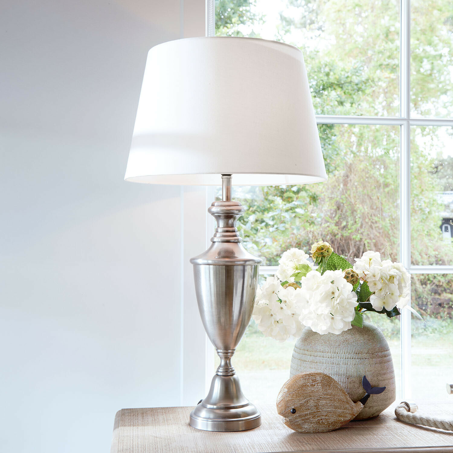 Lampa stołowa Montalure Lampa stołowa z metalową podstawą i białym abażurem na drewnianym stole z dekoracją kwiatową