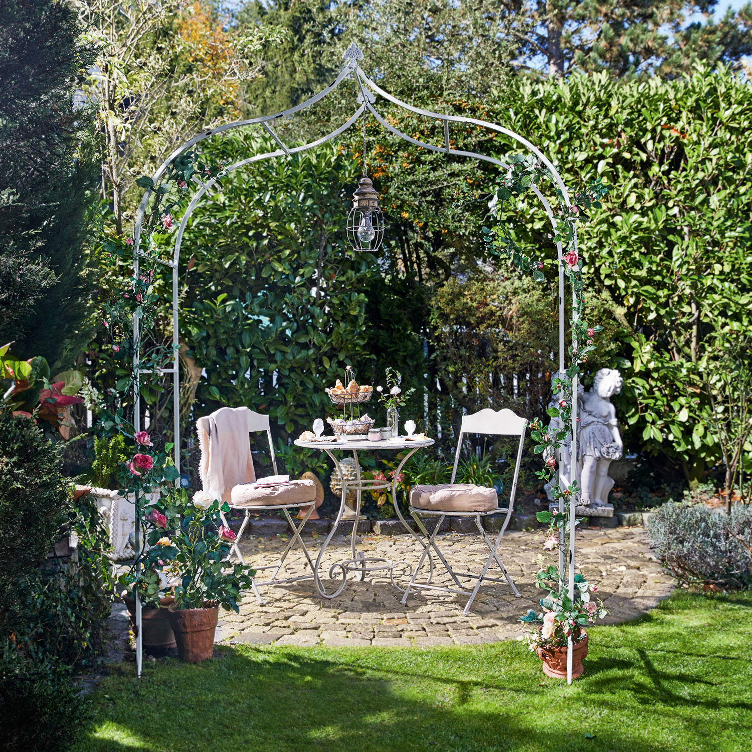 Pergola ogrodowa Dimarus Biały metalowy łuk ogrodowy z różowymi kwiatami i lampą, przy kamiennym patio z meblami