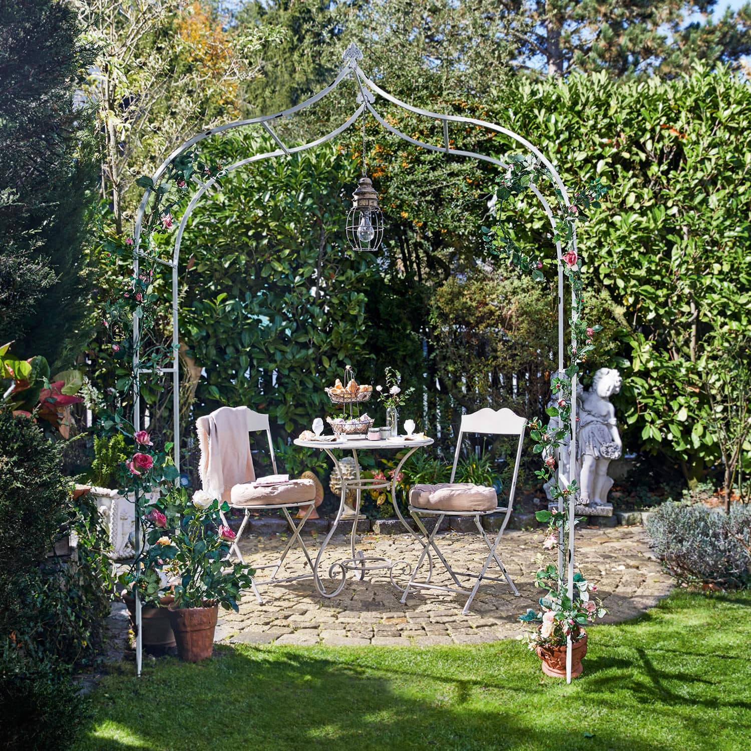 Pergola ogrodowa Dimarus Biały metalowy łuk ogrodowy z różowymi kwiatami i lampą, przy kamiennym patio z meblami