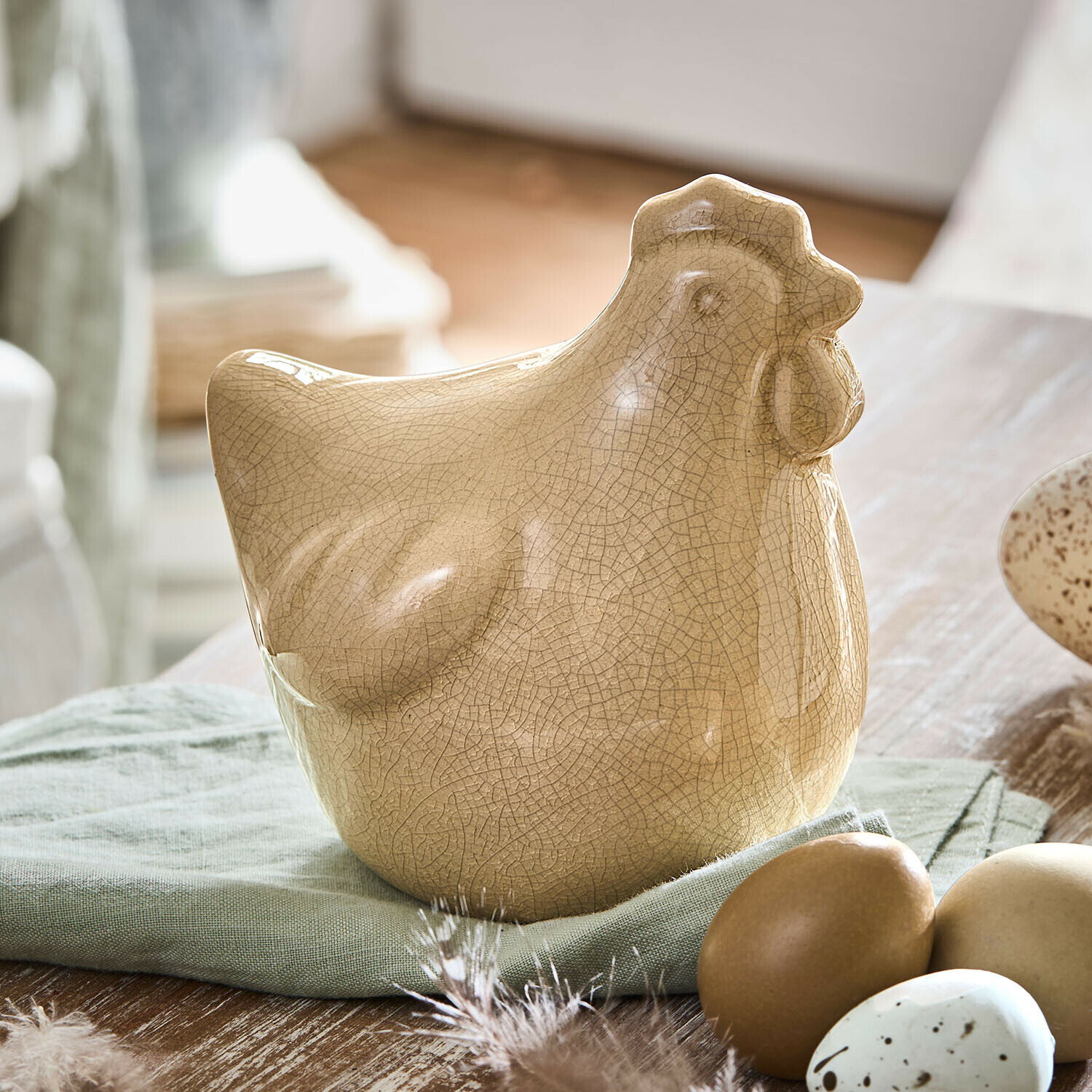 Ozdobna kura Roisenn Beżowa ceramiczna figurka kury z efektem spękań na zielonej tkaninie obok dekoracyjnych jajek