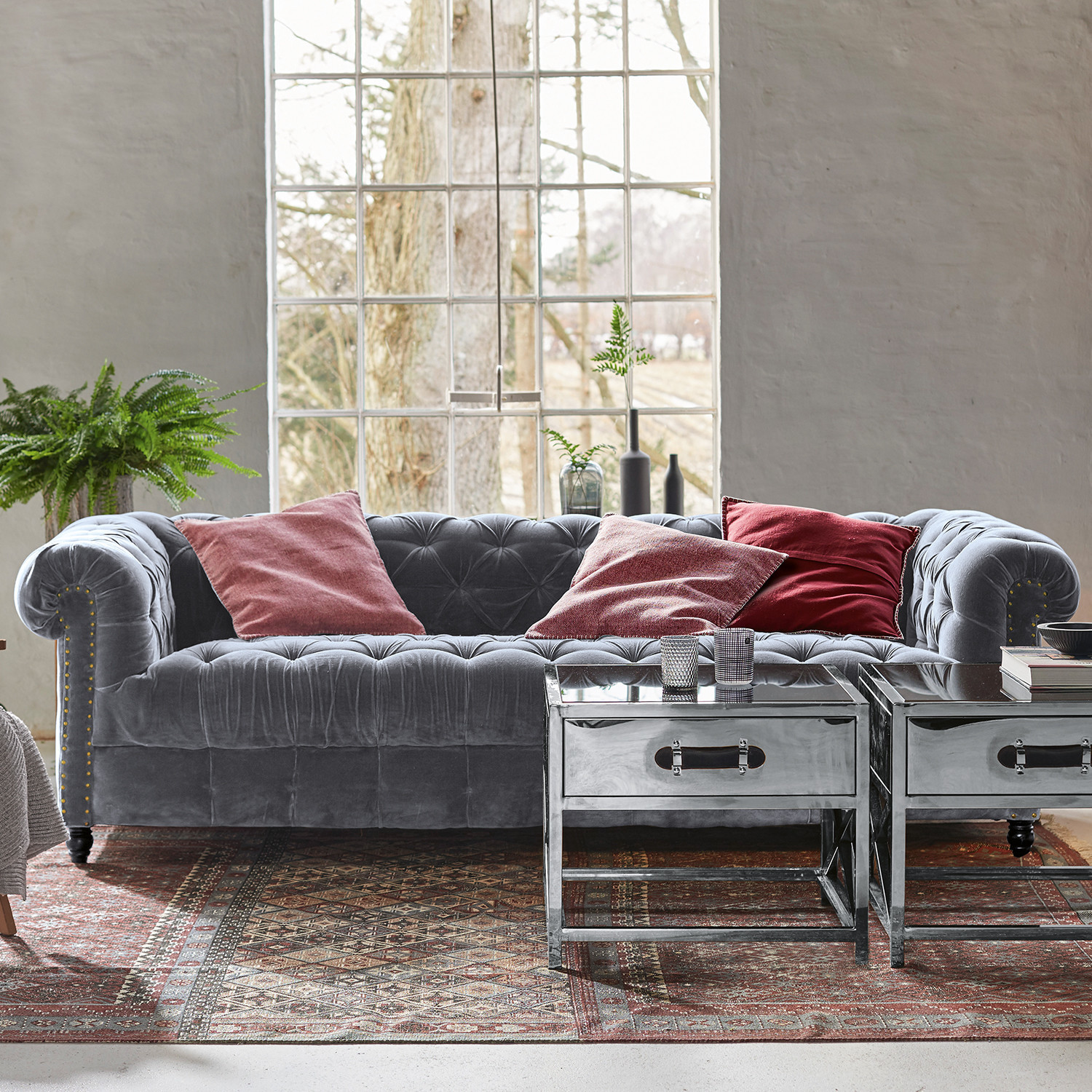 Sofa Parpeville Szara aksamitna sofa Chesterfield z czerwonymi poduszkami i metalowymi stolikami kawowymi na dywanie