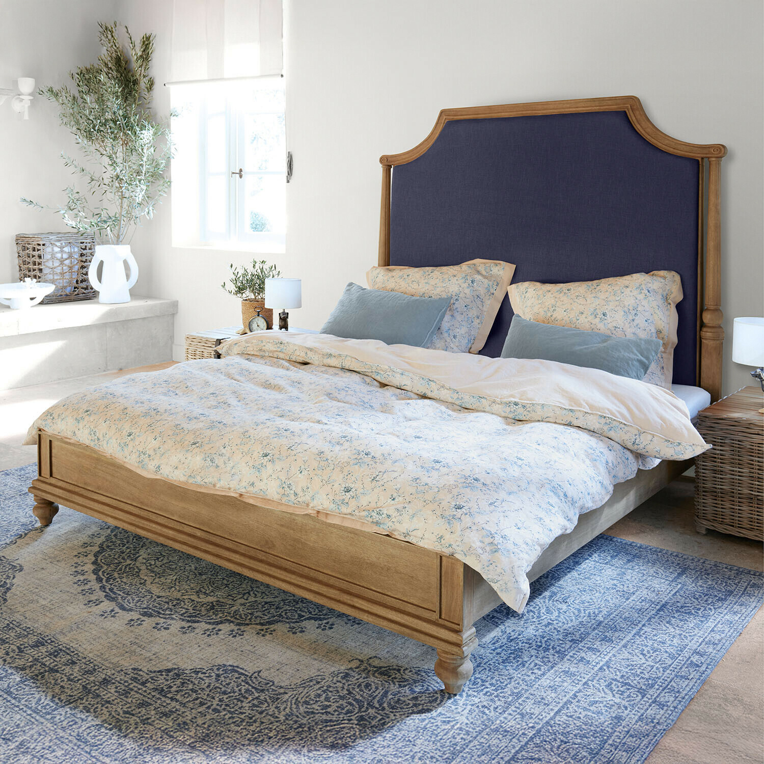 Łóżko z drewnianą ramą i granatowym tapicerowanym zagłówkiem w jasnej sypialni z niebieskim dywanem