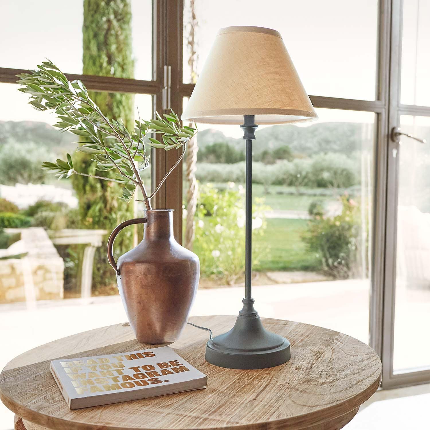 Metalowy dzbanek w kolorze miedzi z gałązką i lampa stołowa z beżowym abażurem na drewnianym stole