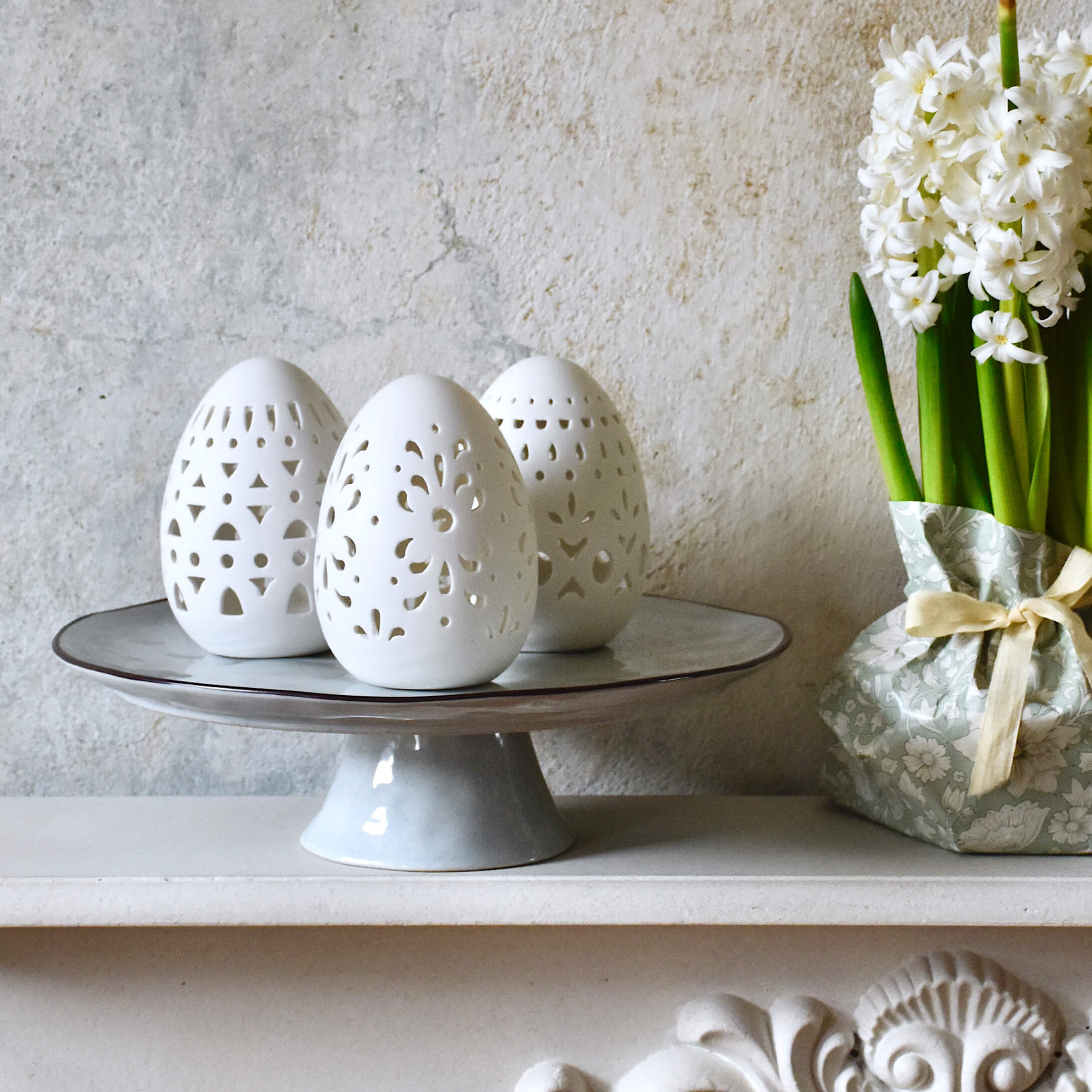 Białe ceramiczne pisanki z ażurowym wzorem na szarej paterze obok białych kwiatów w ozdobnym opakowaniu