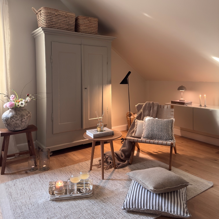 Przytulny salon z beżowym fotelem, drewnianym stolikiem i szafką pod skosem, dekoracje i lampy