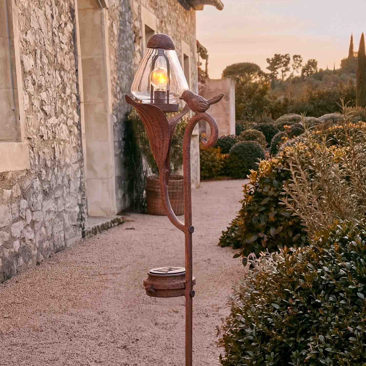 Ozdobna lampa ogrodowa z metalu w kolorze rdzy z motywem ptaka i szklanym kloszem na ścieżce