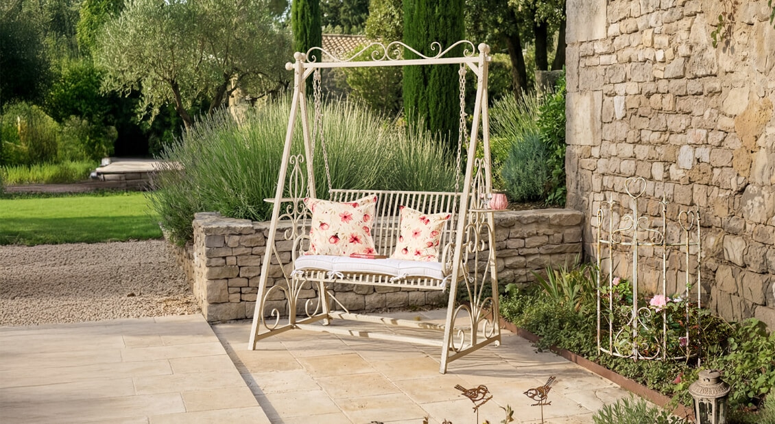 Biała metalowa huśtawka ogrodowa z czerwono-białymi poduszkami w kwiaty na tarasie z kamienia