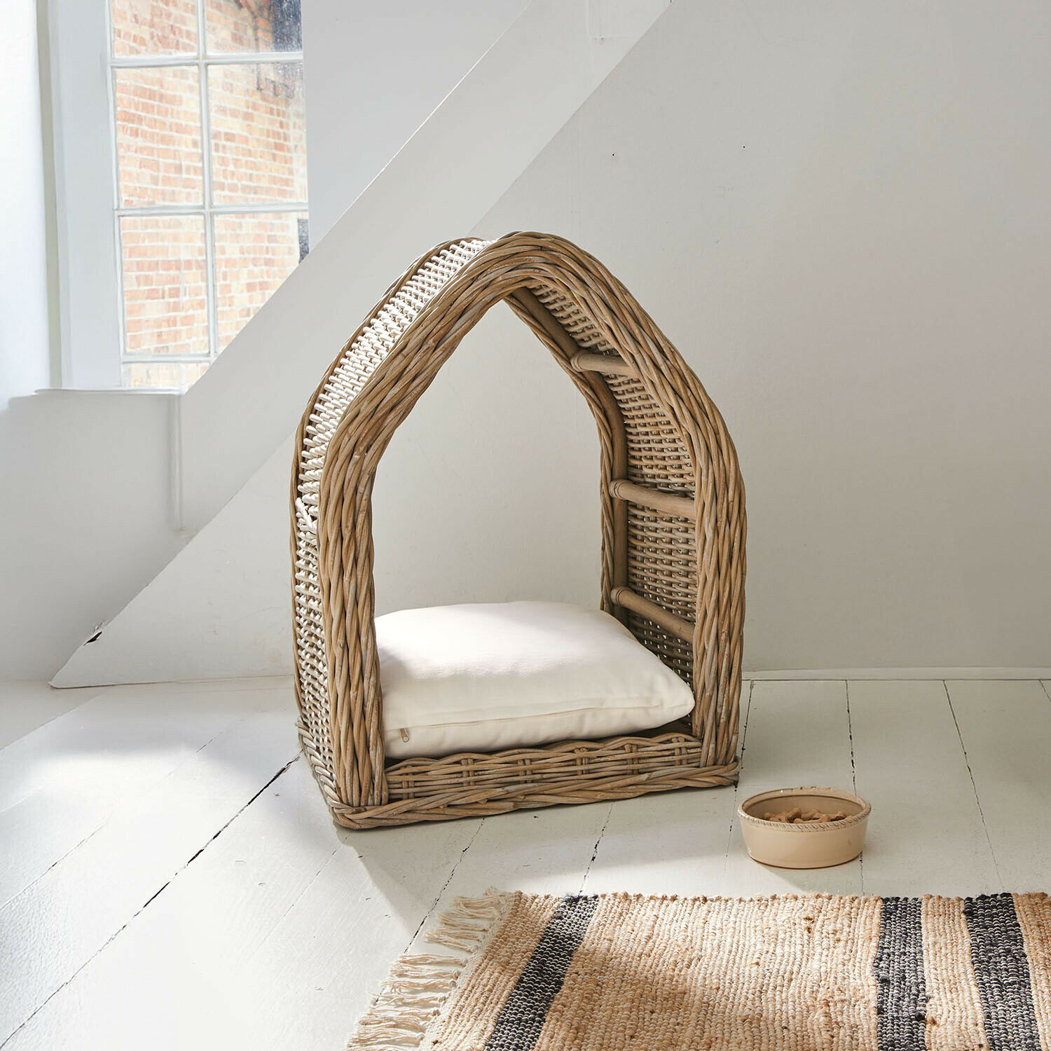 Domek dla kota z rattanu w naturalnym kolorze z białą poduszką i miską na przysmaki