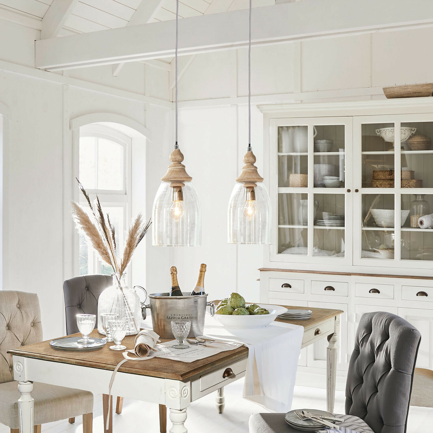Jadalnia w stylu rustykalnym z białym drewnianym stołem i szarymi tapicerowanymi krzesłami oraz szafką z przeszklonymi drzwiami