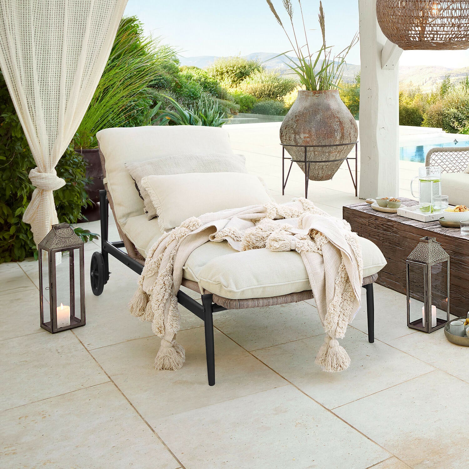 Leżak ogrodowy z kremowymi poduszkami i miękkim kocem na tarasie z widokiem na basen i zieleń