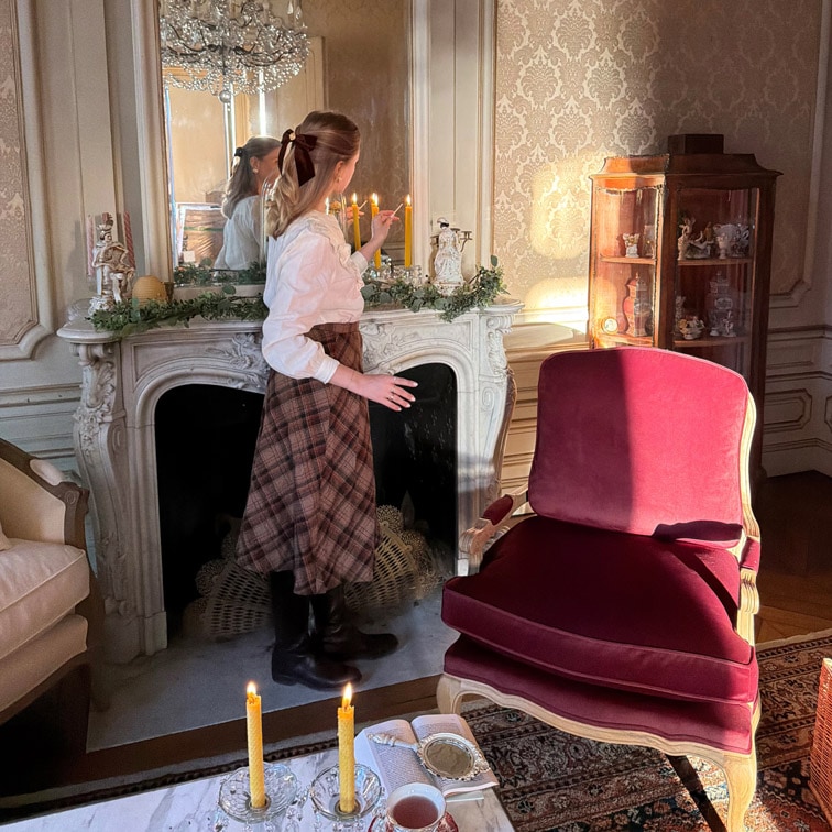 Kobieta zapalająca świece przy marmurowym kominku w eleganckim salonie z bordowym fotelem