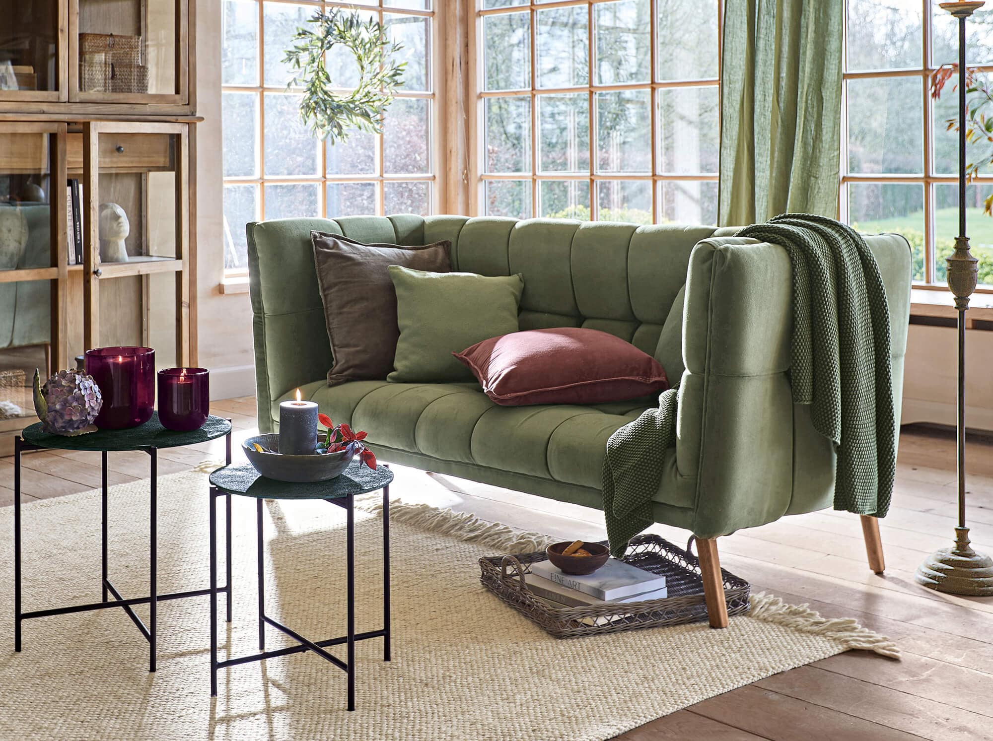 Zielona aksamitna sofa vintage z poduszkami i pledem w jasnym salonie z dużymi oknami
