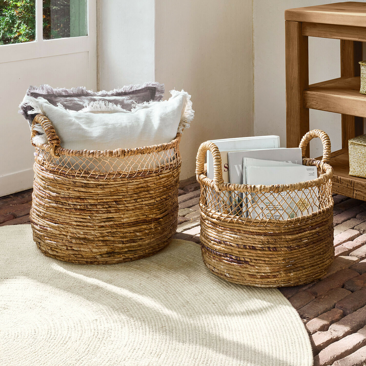 Dwa wiklinowe kosze do przechowywania w naturalnym kolorze z uchwytami, wypełnione poduszkami i książkami