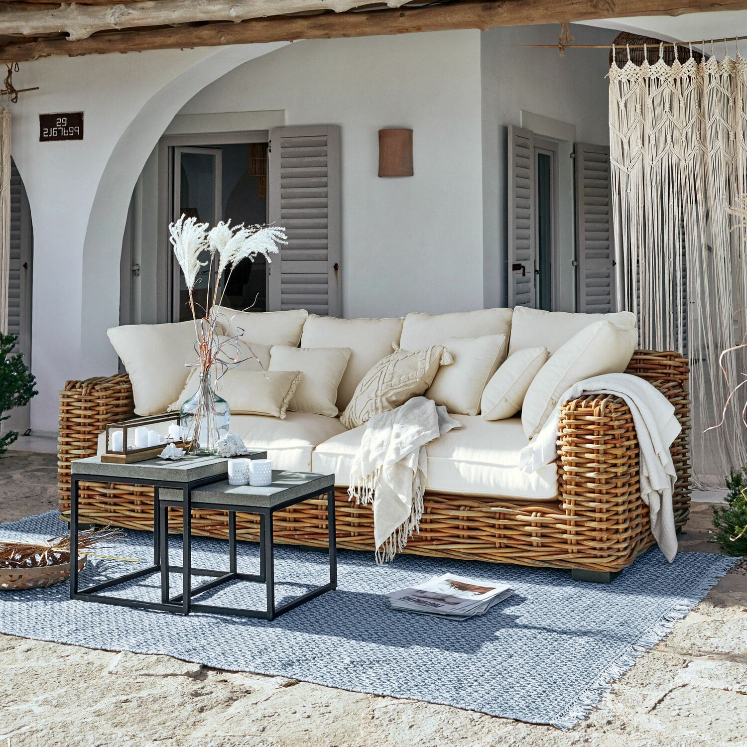 Sofa Rimini Sofa z rattanu z kremowymi poduszkami i kocem na niebieskim dywanie na tarasie