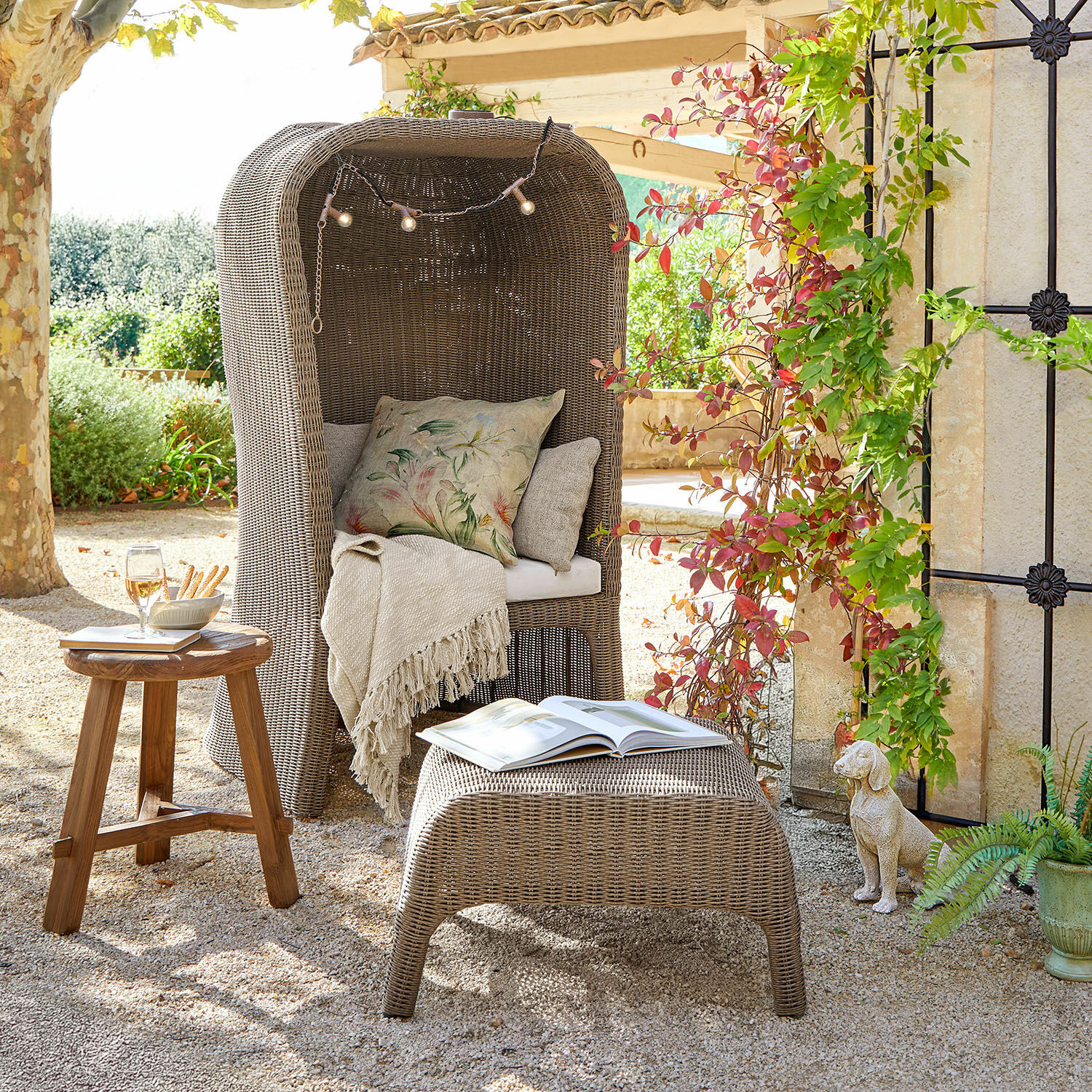 Szary fotel z rattanu z poduszkami i pledem na zewnątrz, stolik drewniany i pufa z książką