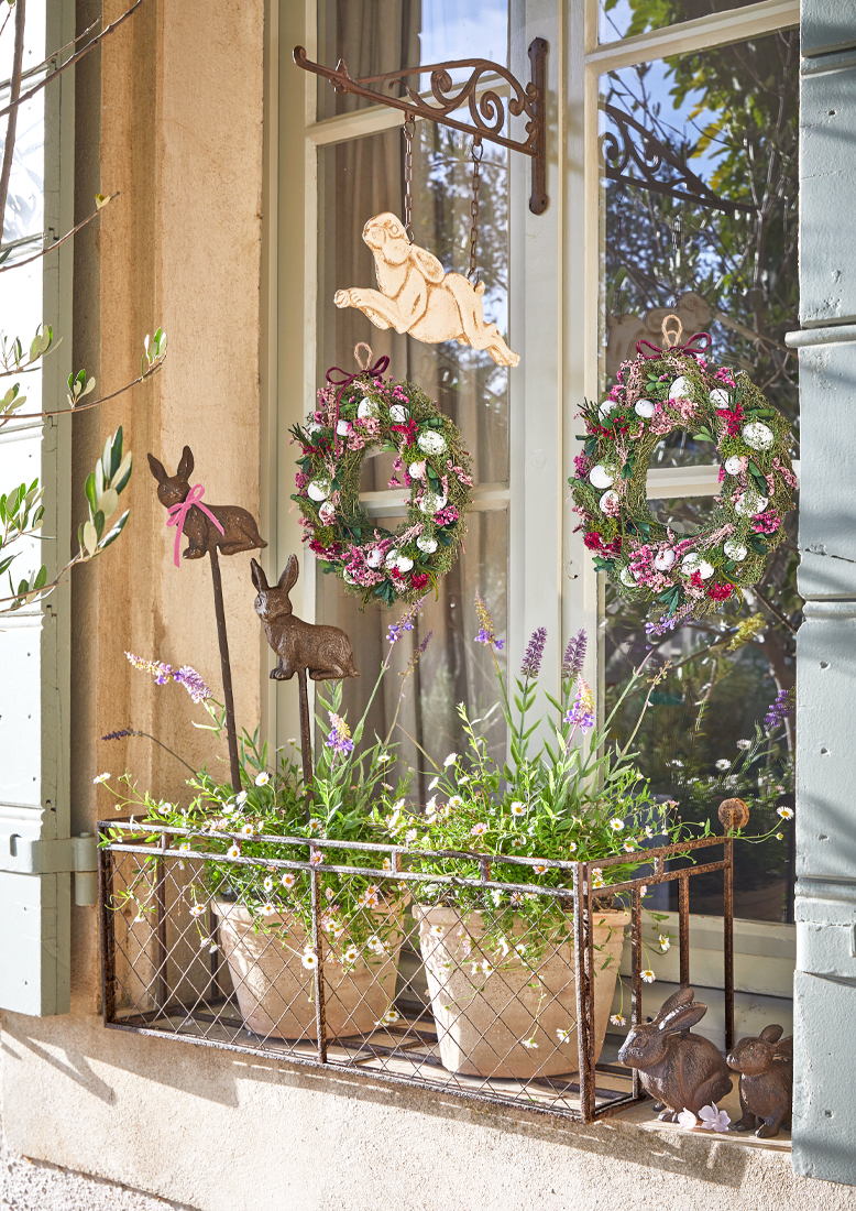 Dekoracja okienna z metalowym stojakiem, donicami z kwiatami i wiosennymi wieńcami z jajkami i królikami