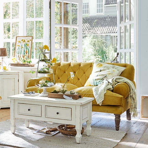 Żółta aksamitna sofa w stylu vintage w jasnym salonie z białą drewnianą ławą i dekoracjami