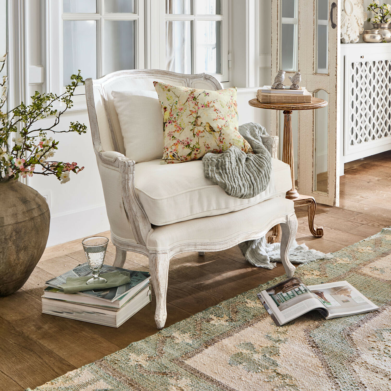 Fotel Kingswinford Biały drewniany fotel z jasną tapicerką, ozdobną poduszką i szarym pledem w przytulnym salonie