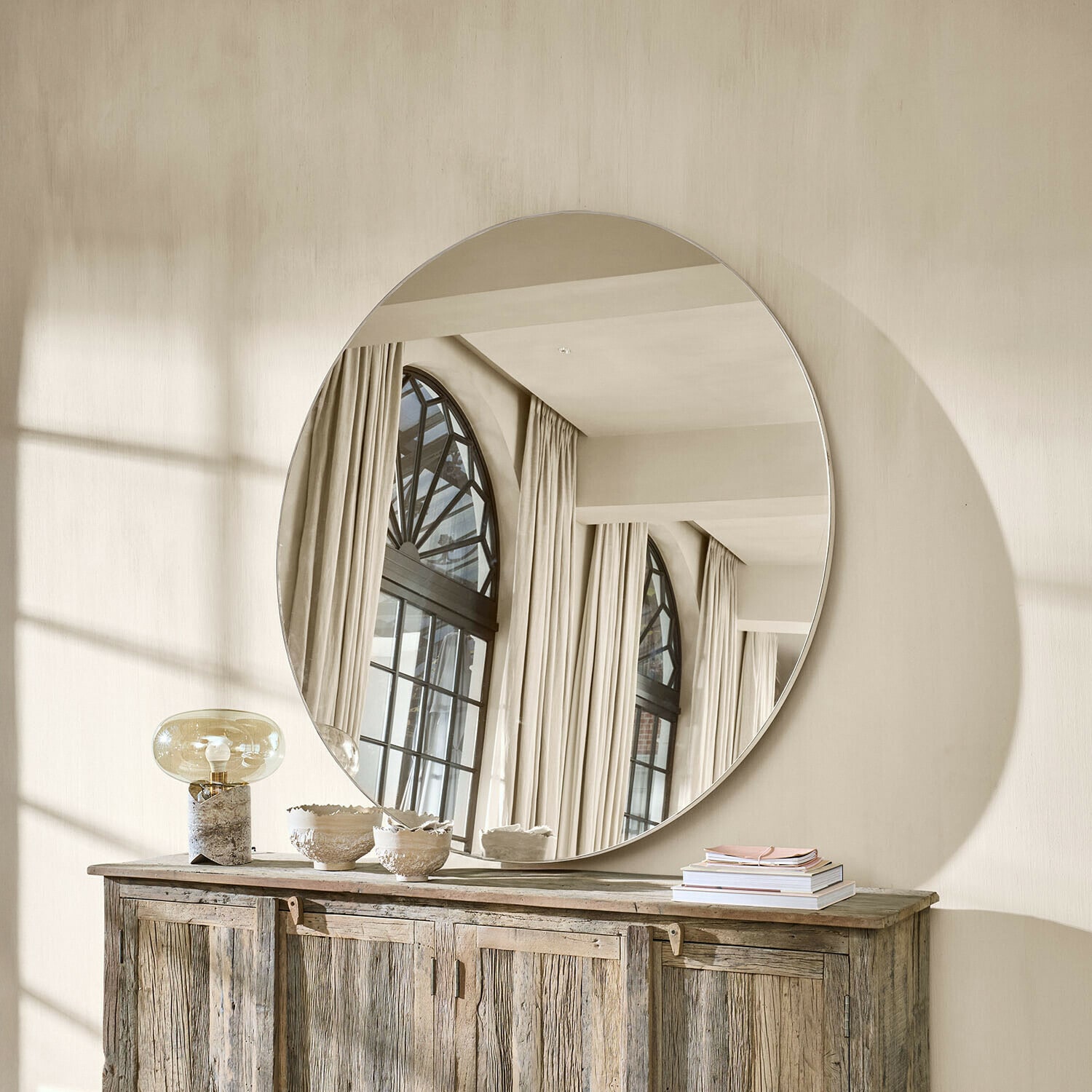 Duże okrągłe lustro na ścianie nad drewnianą komodą w jasnym, naturalnym wnętrzu