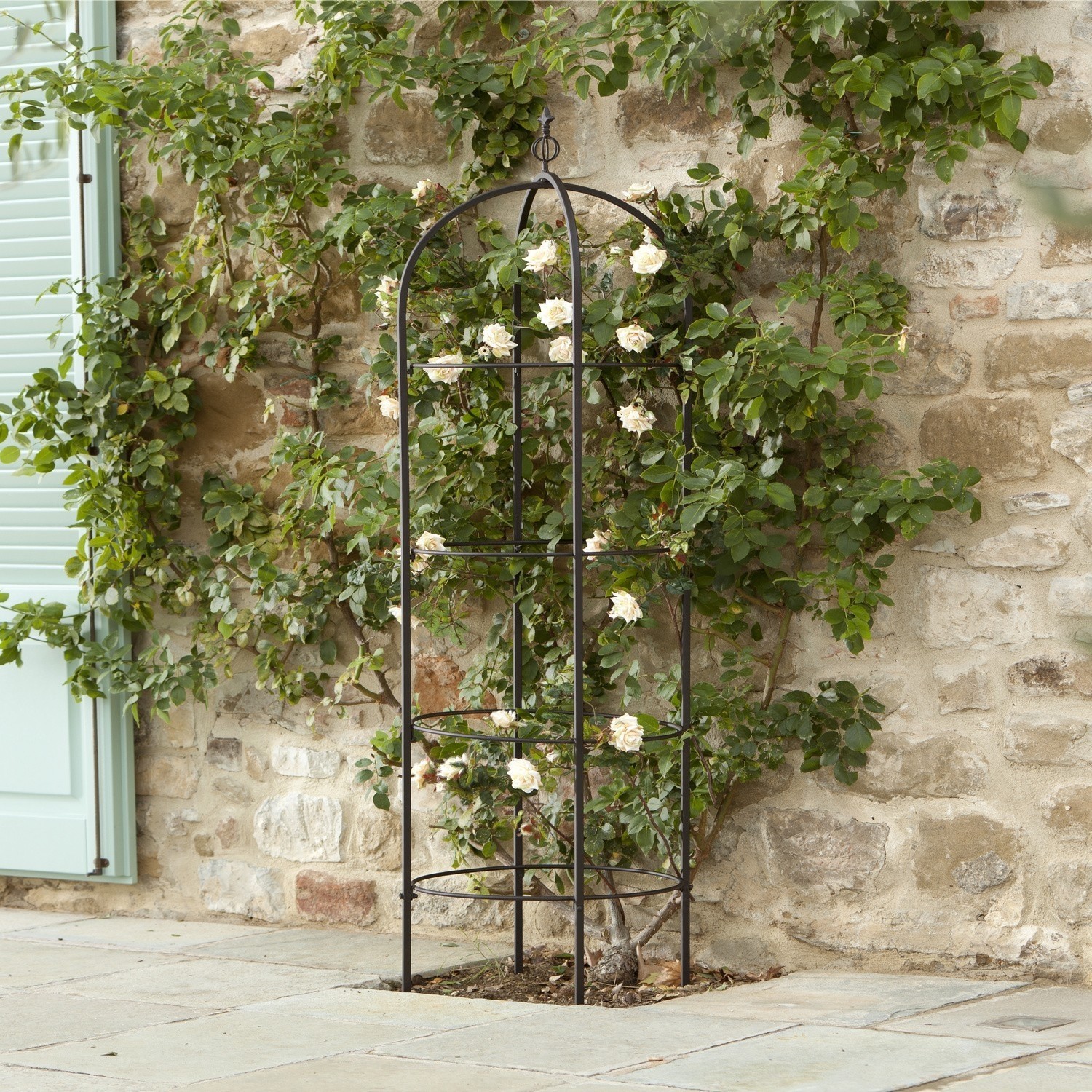 Obelisk ogrodowy Besson Metalowa czarna kratownica ogrodowa do podpierania pnączy na tle kamiennej ściany i zielonych liści