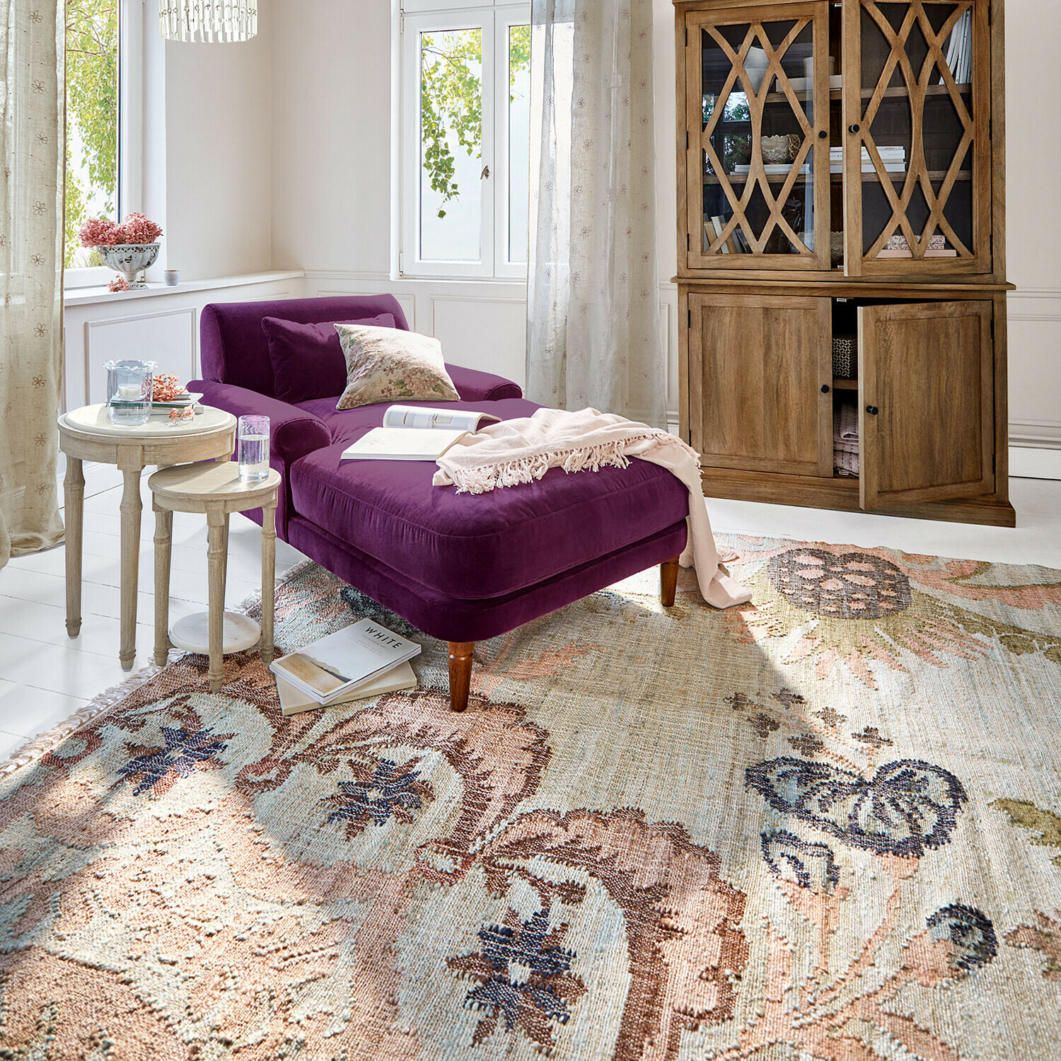 Fioletowa aksamitna leżanka w jasnym salonie z drewnianą szafką i dekoracyjnym dywanem