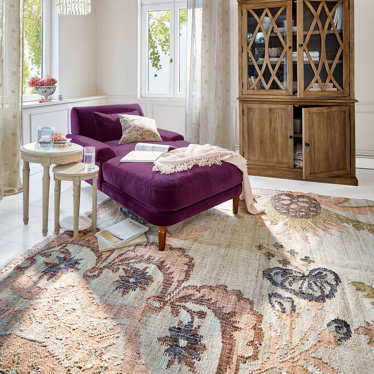 Fioletowa aksamitna leżanka w jasnym salonie z drewnianą witryną i ozdobnym dywanem