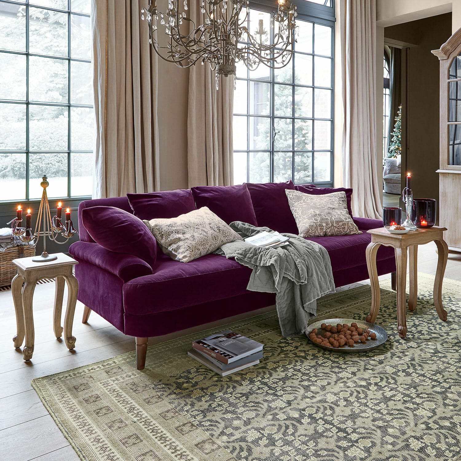 Sofa Rivermere Fioletowa welurowa sofa z poduszkami i szarym kocem w jasnym salonie z dużymi oknami i drewnianymi stolikami