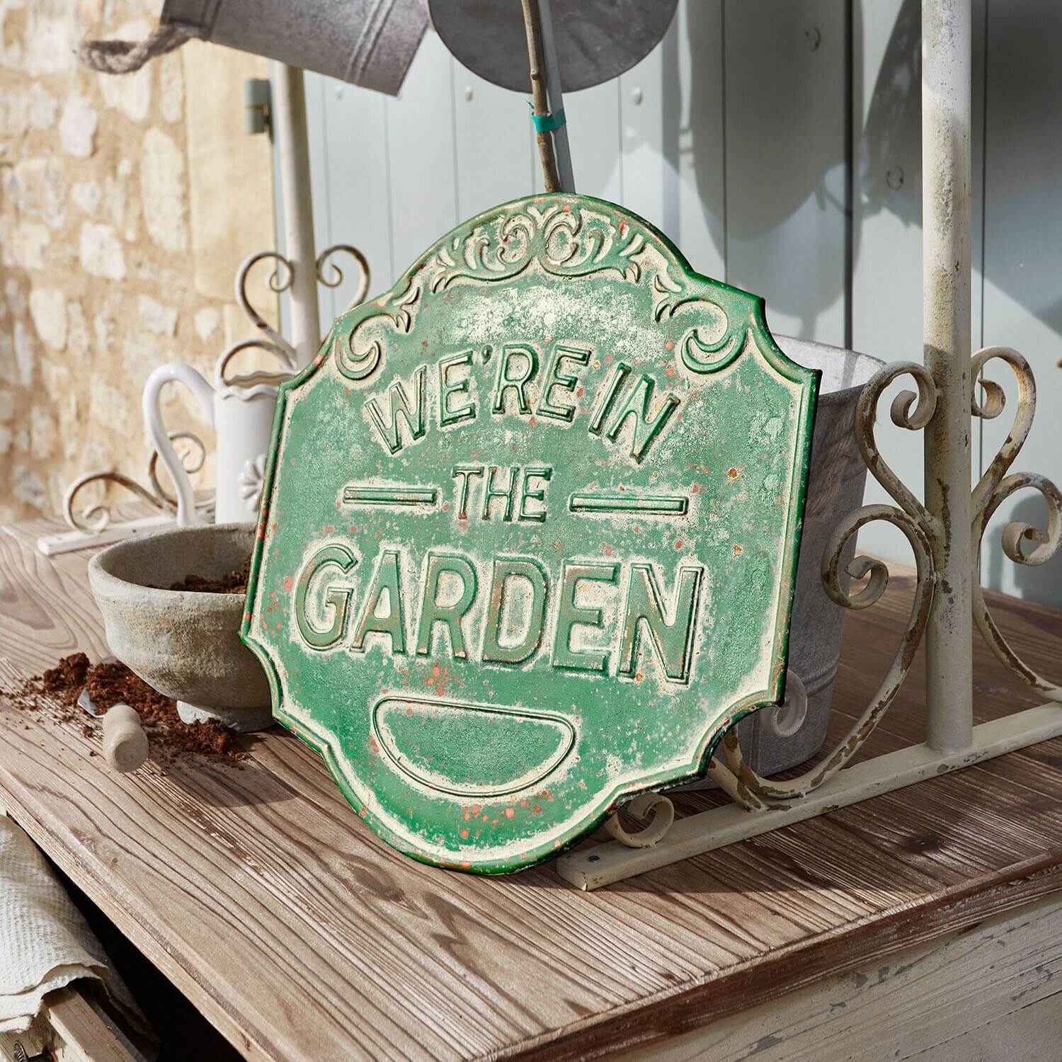 Tabliczka dekoracyjna Merrin Zielona metalowa tablica dekoracyjna z napisem We're in the Garden na drewnianym stole w ogrodzie