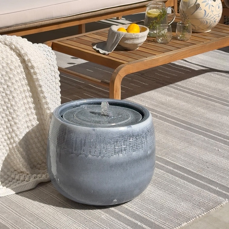 Szara ceramiczna fontanna stołowa na jasnym dywanie obok drewnianego stolika i białego koca