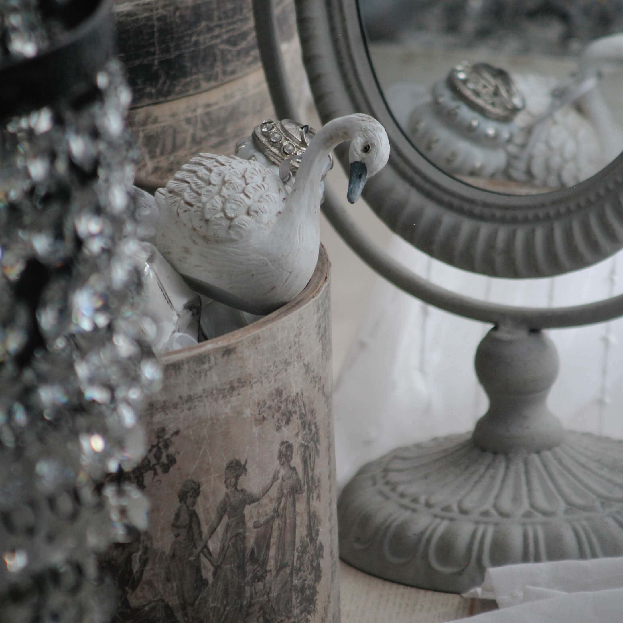 Biała ceramiczna figurka łabędzia obok ozdobnego lustra i vintage pojemnika z klasycznym wzorem