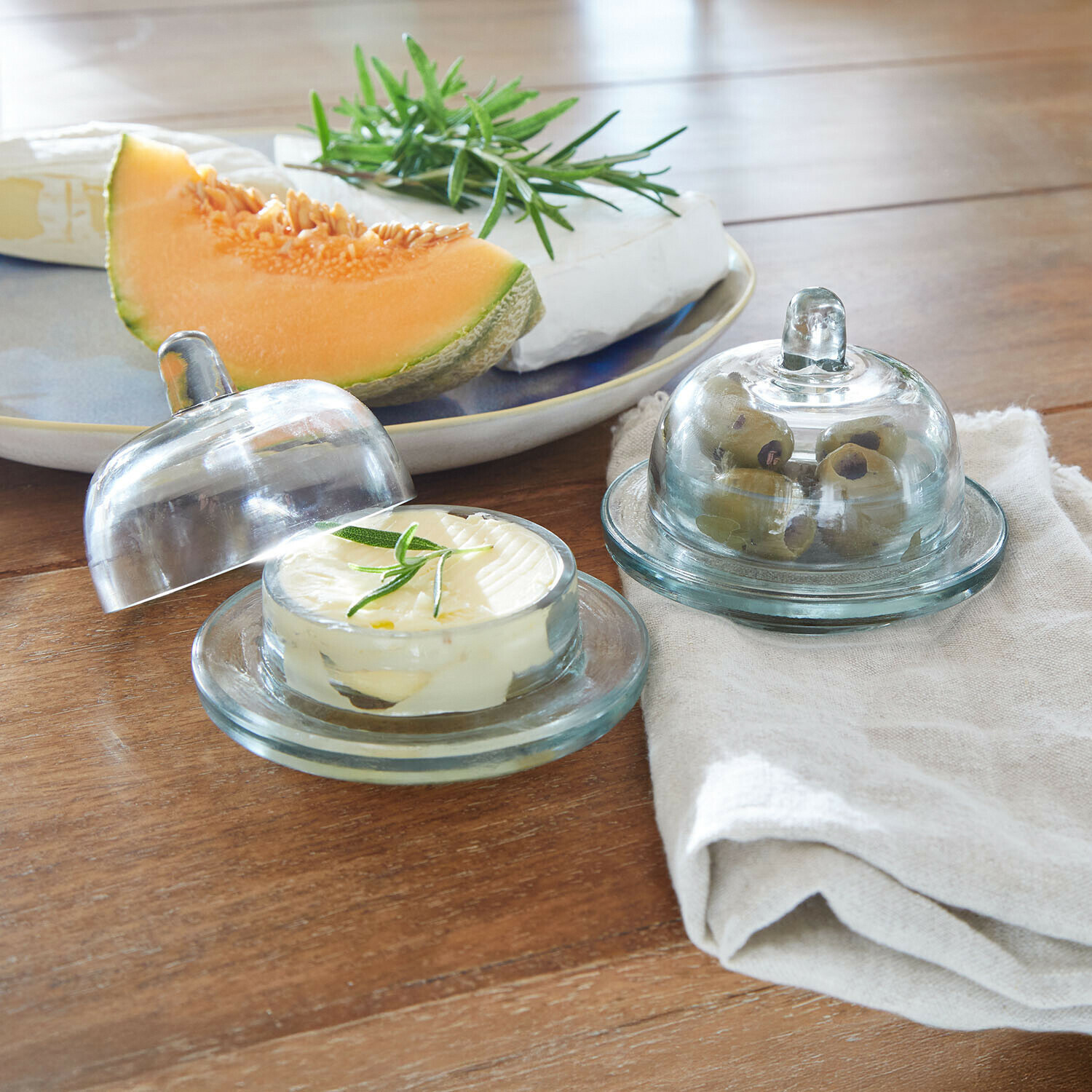 Szklane pojemniki na masło i oliwki na drewnianym stole z melonem i serem w tle