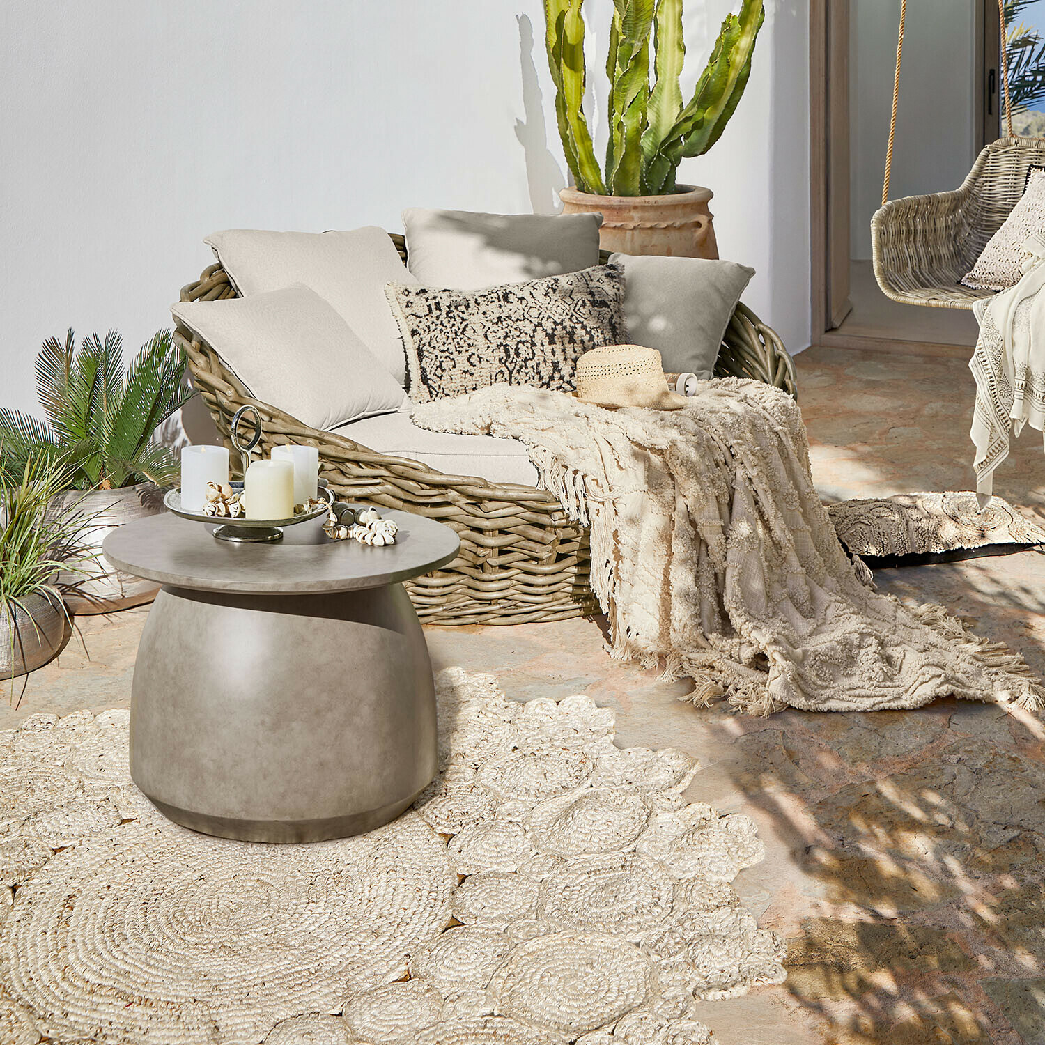 Owalna sofa z rattanu z kremowymi poduszkami i pledem na tarasie z betonowym stolikiem