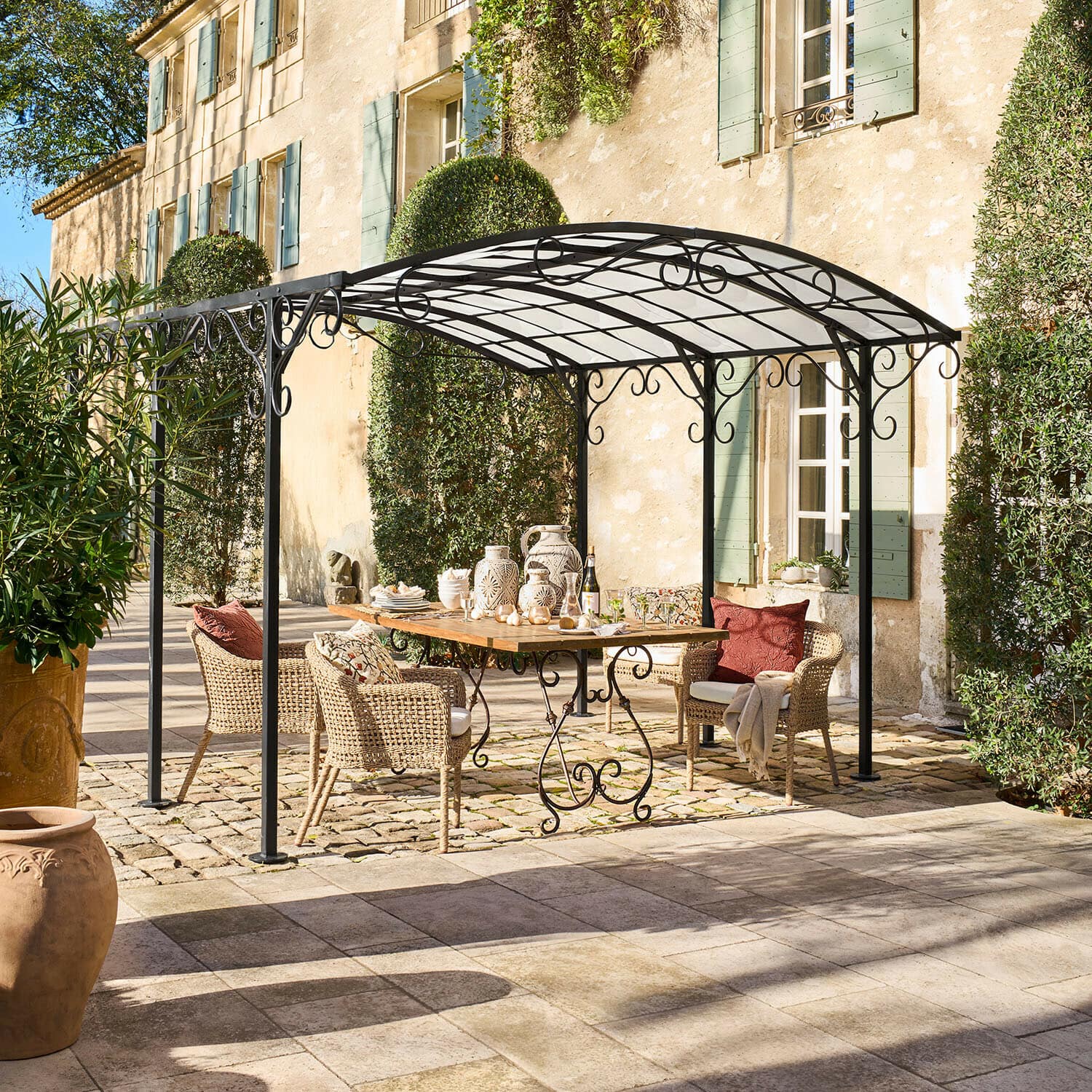 Pergola Delbridge Altana ogrodowa z czarnego metalu z ozdobnymi zawijasami nad drewnianym stołem i wiklinowymi krzesłami