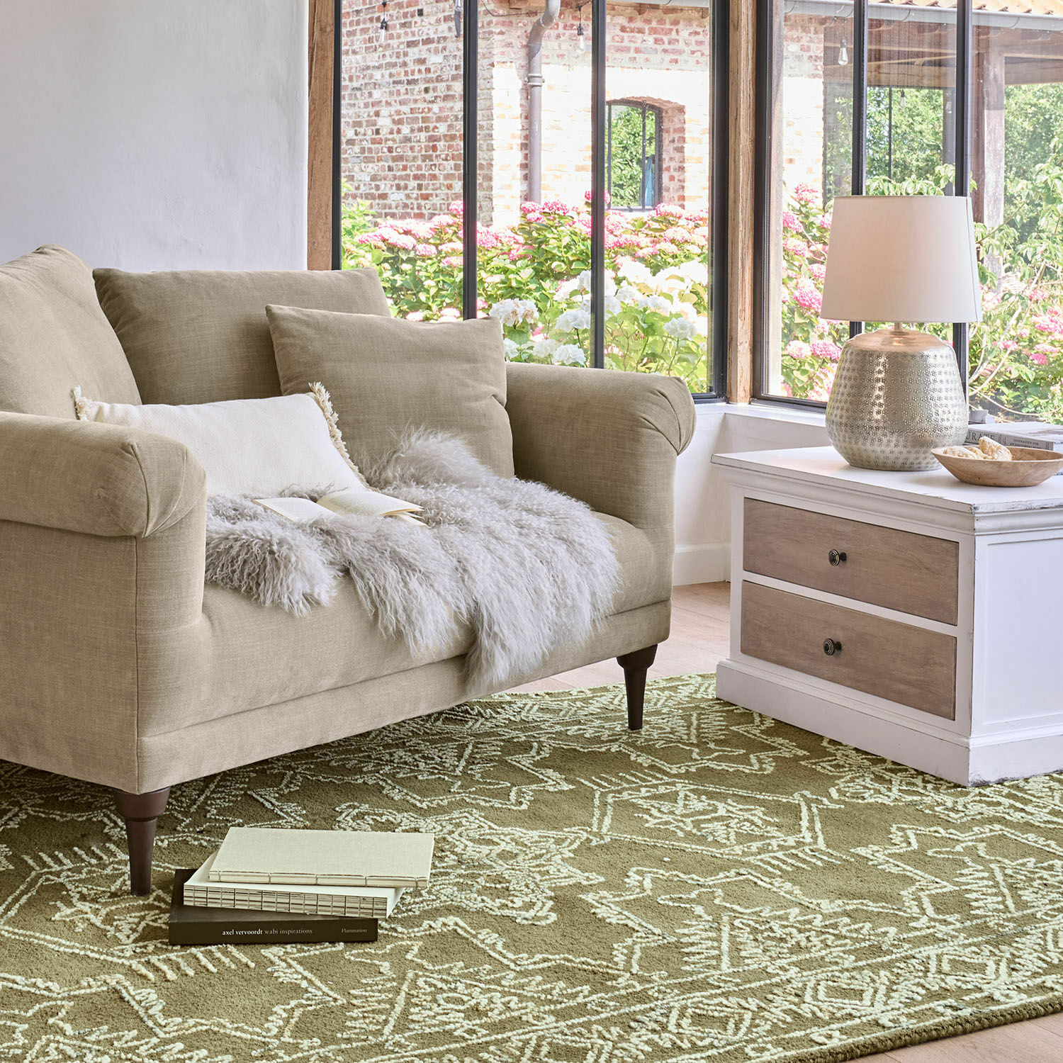Dywan Qazoura Beżowa sofa z tkaniny z poduszkami i futrzanym kocem na zielonym dywanie z geometrycznym wzorem