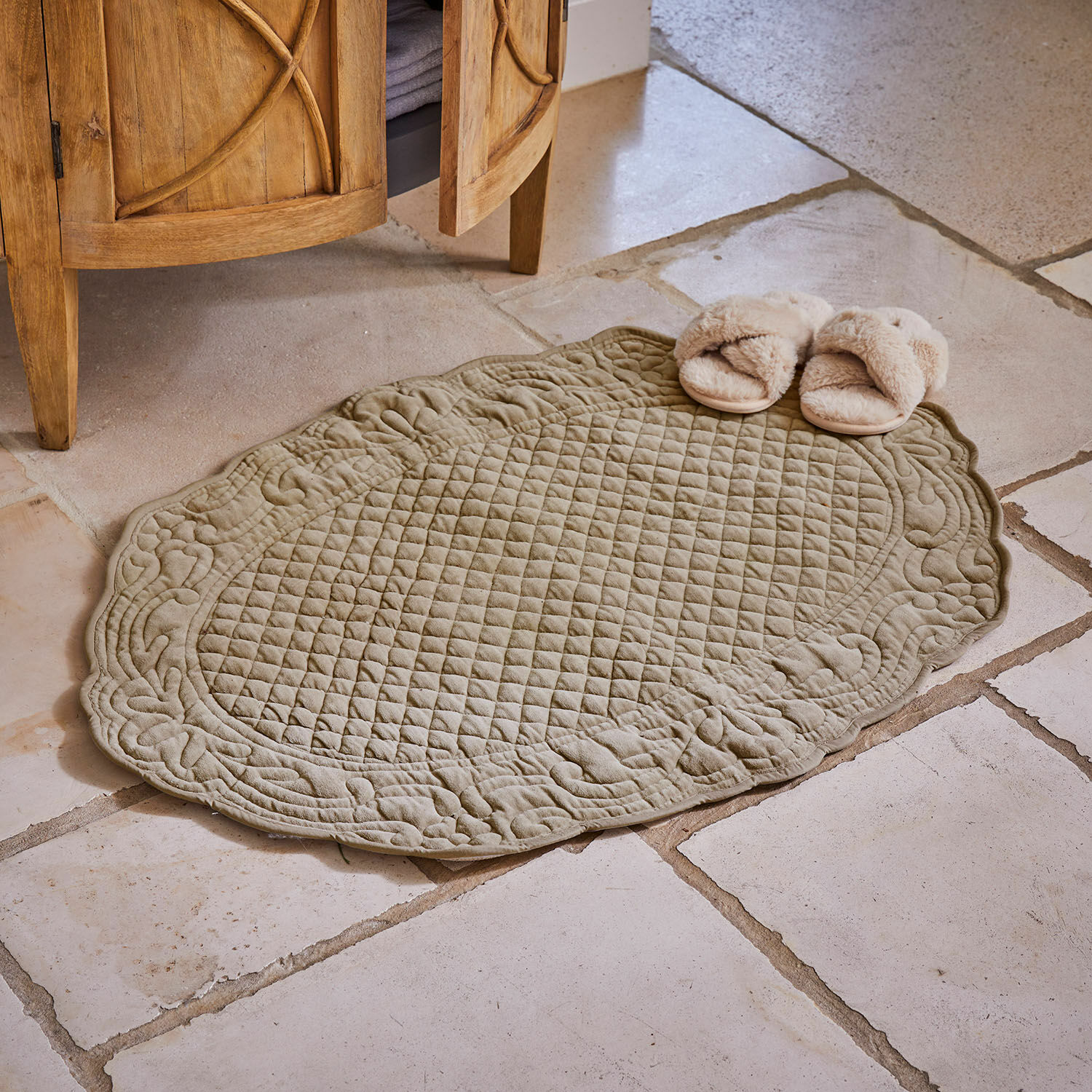Owalny, pikowany dywanik kąpielowy w kolorze oliwkowym na kamiennej podłodze z beżowymi kapciami