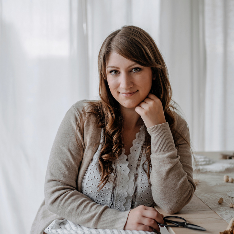 Portret kobiety w beżowym swetrze i białej koronkowej bluzce przy stole w jasnym wnętrzu
