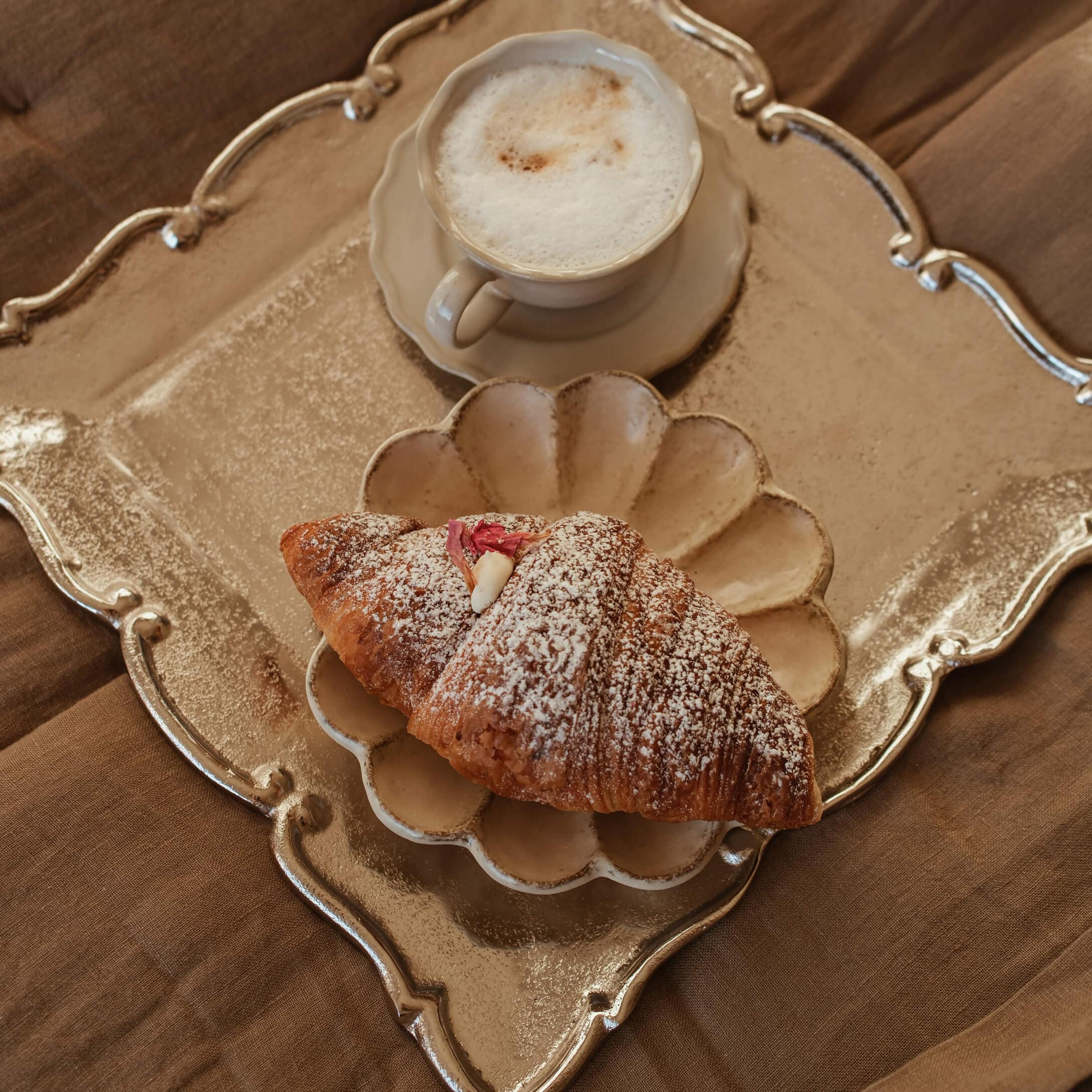 Kremowa filiżanka cappuccino i croissant z cukrem pudrem na beżowym talerzu i złotej tacy