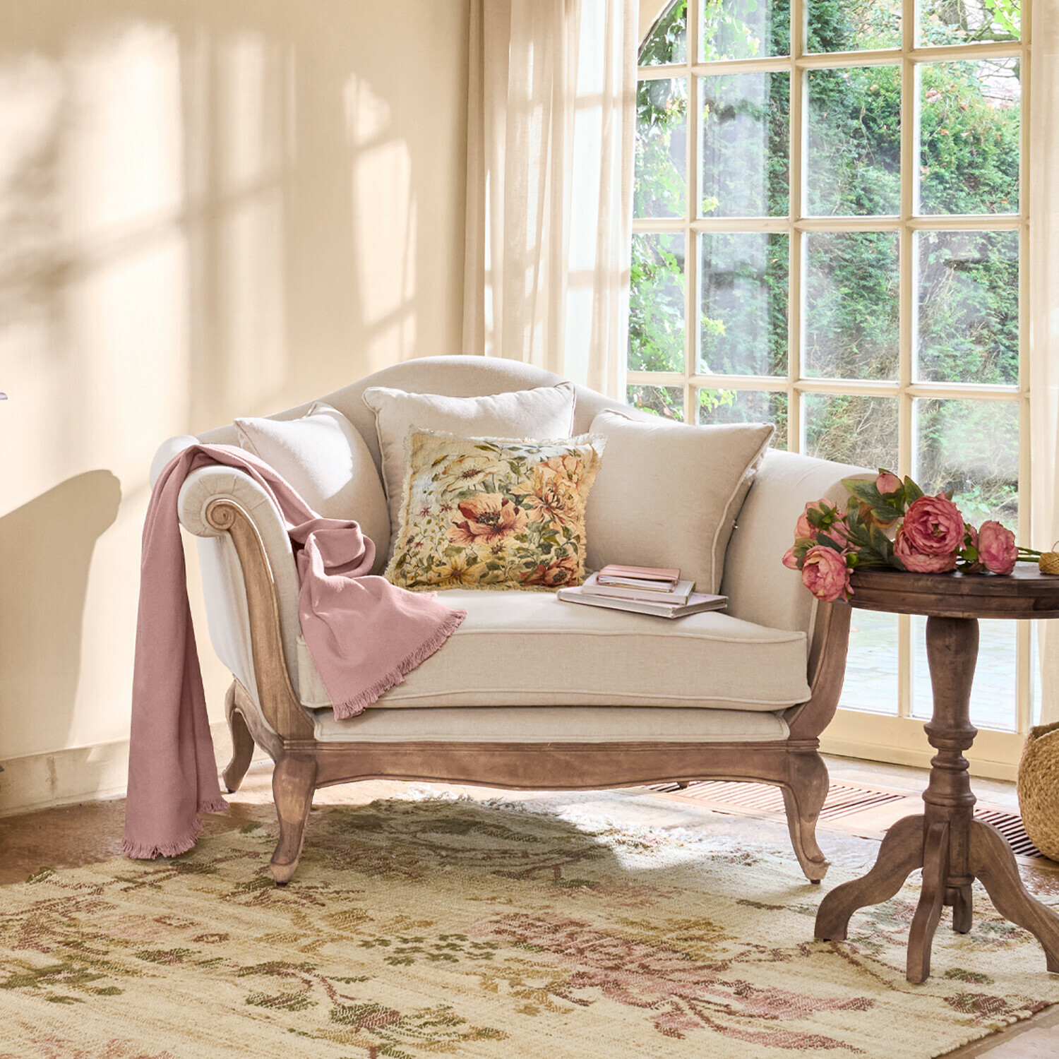 Beżowa sofa z drewnianą ramą, ozdobiona poduszką w kwiaty i różowym kocem w jasnym salonie