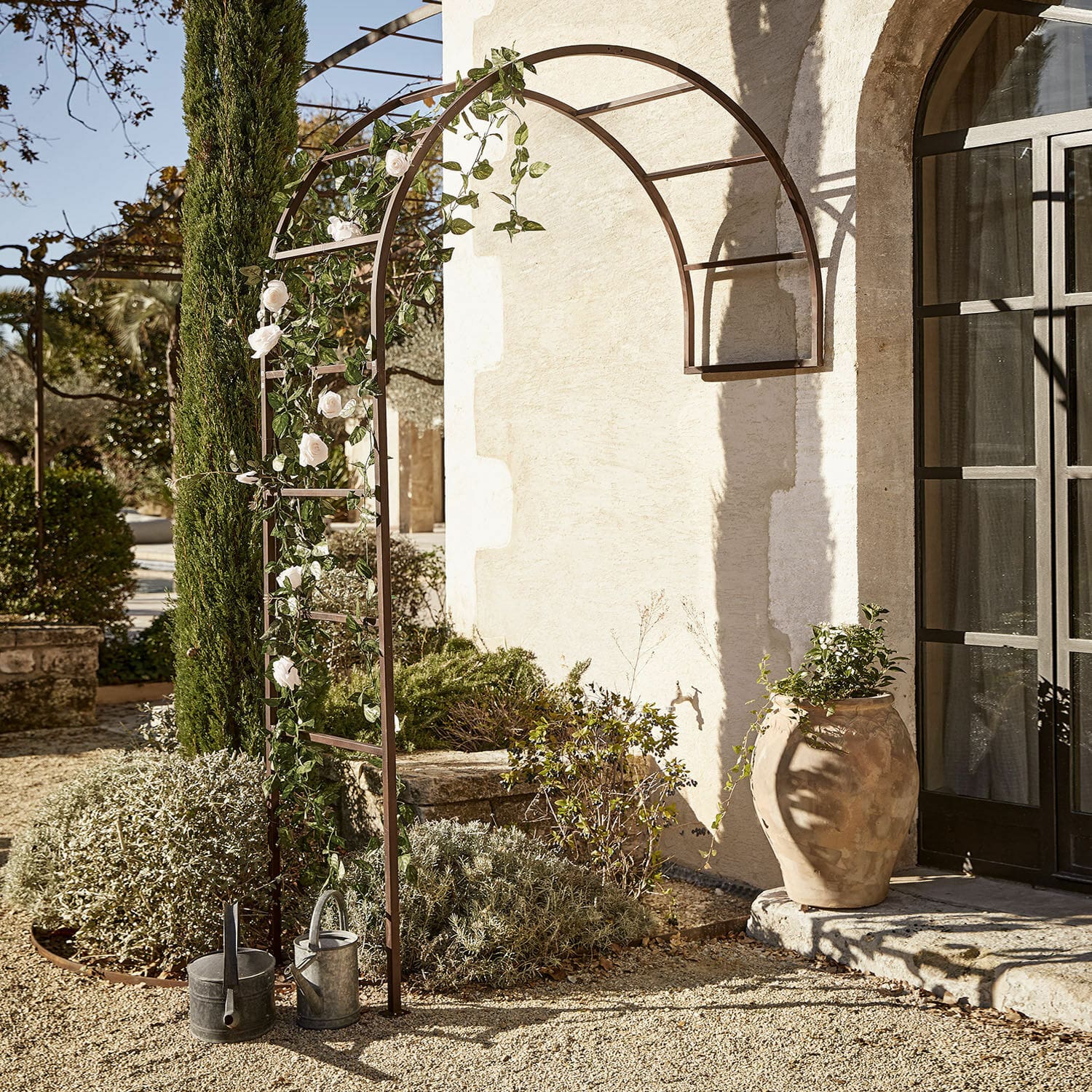 Pergola ogrodowa Auburn Metalowa łukowa pergola ogrodowa w kolorze brązowym z dekoracyjnymi różami i konewkami na żwirze