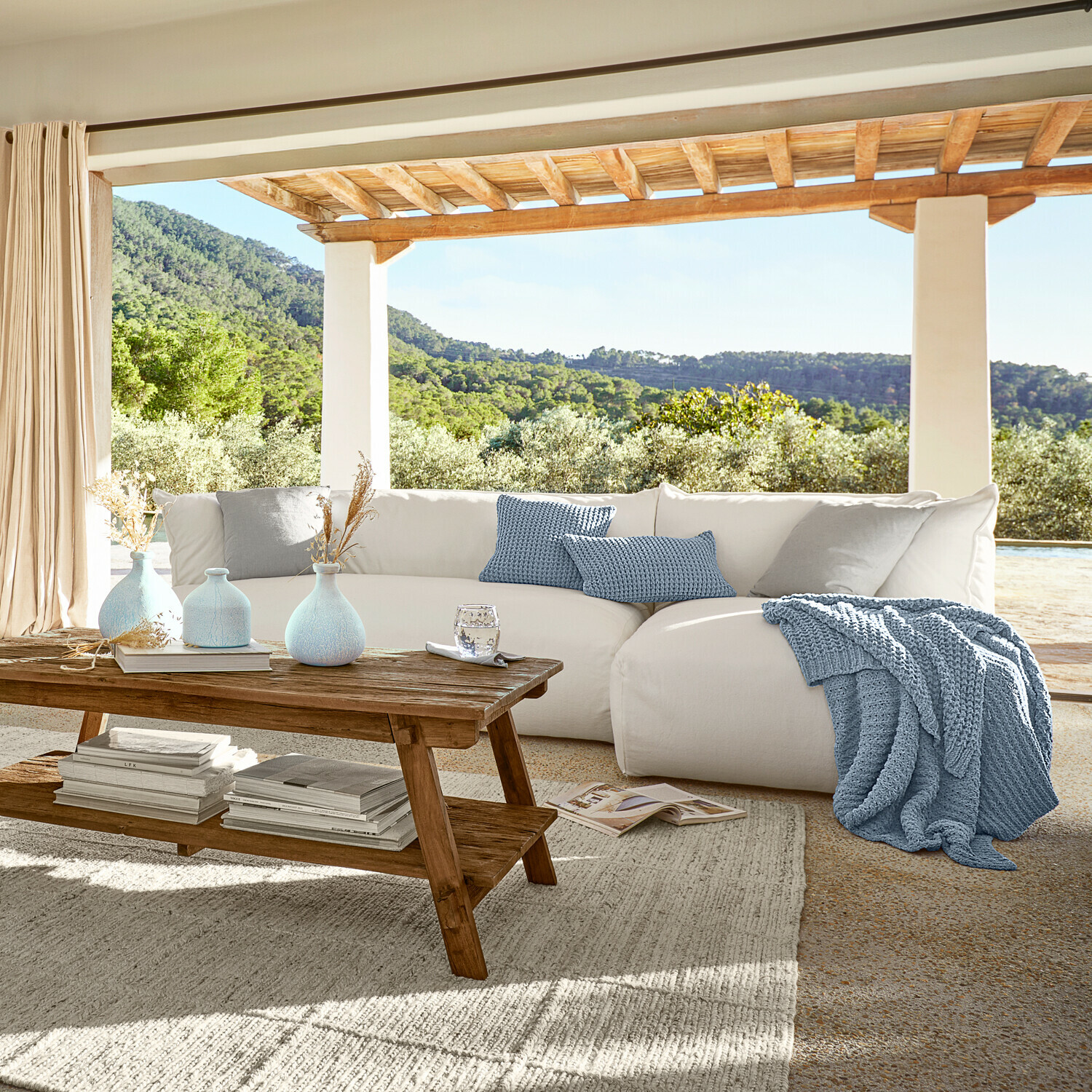 Przytulna biała sofa z niebieskim dzianym kocem i poduszkami na tarasie z widokiem na zieleń