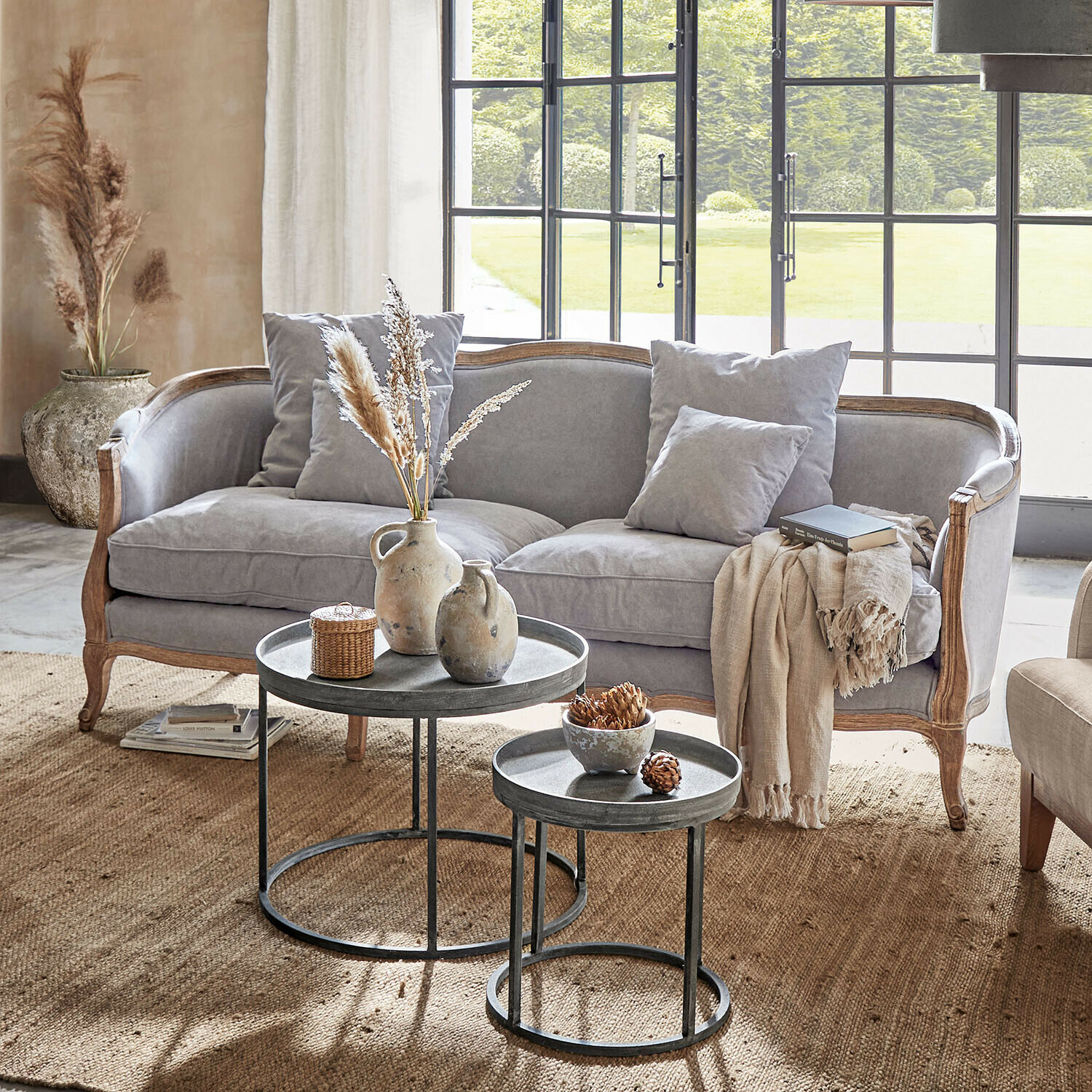 Sofa Monneville Szara sofa z drewnianą ramą i poduszkami w jasnym salonie z metalowymi stolikami kawowymi