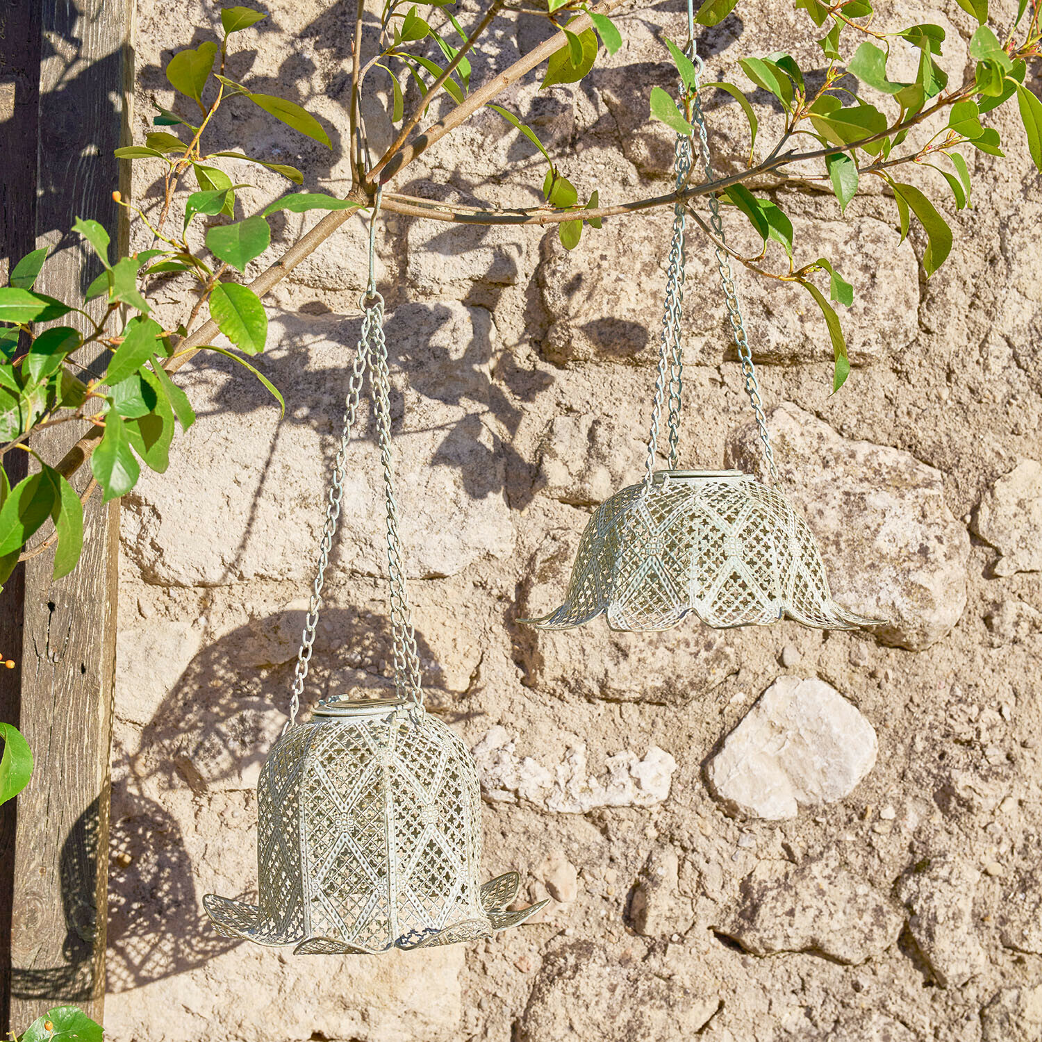 Dwa wiszące metalowe lampiony w kształcie dzwonków w kolorze jasnoszarym na tle kamiennej ściany