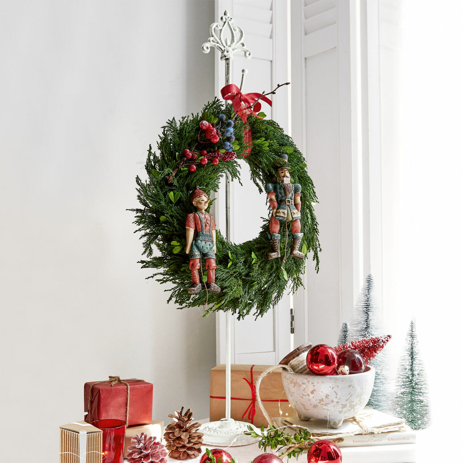 Stojak na wieniec Irulan Świąteczny wieniec z zielonych gałązek z czerwonymi i niebieskimi jagodami oraz drewnianymi figurkami na stojaku