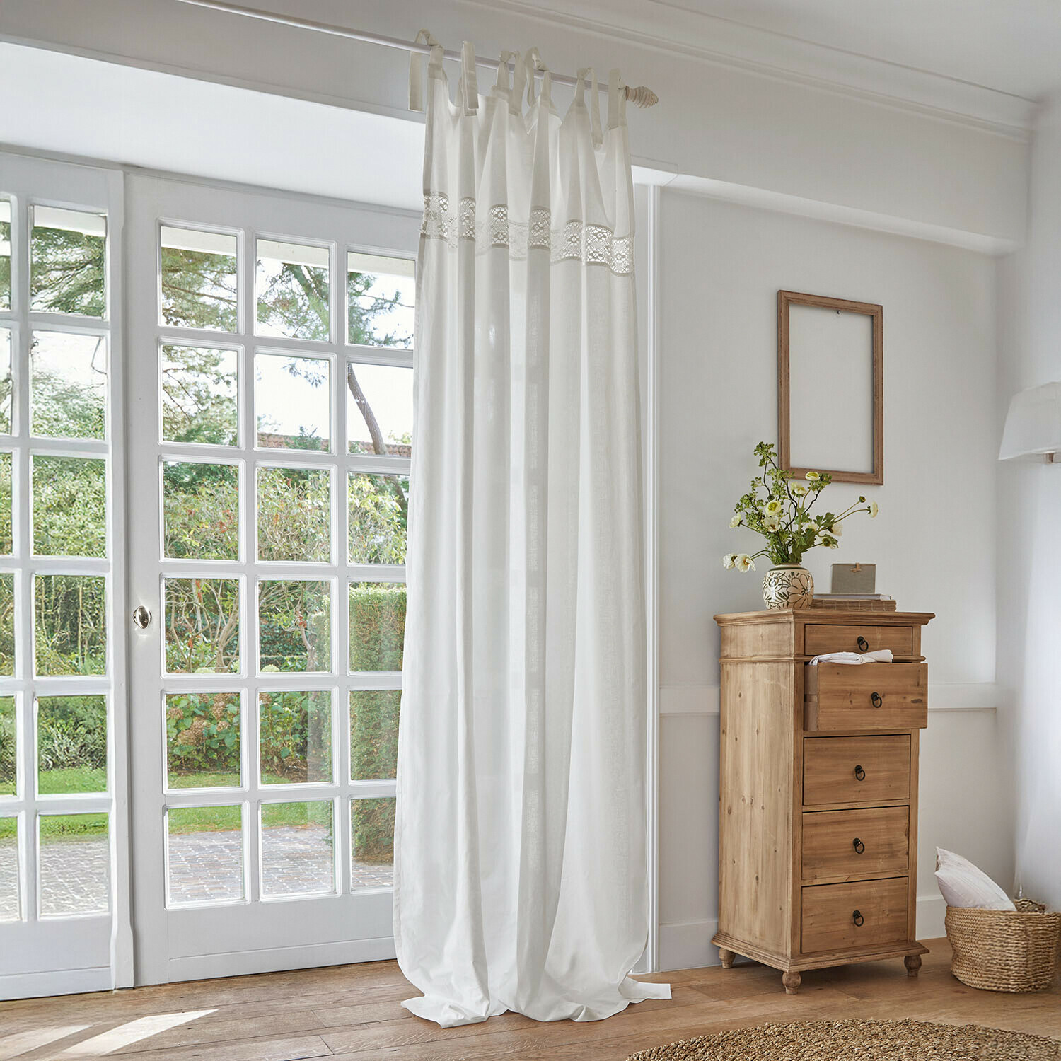 Biała bawełniana zasłona z koronkowym paskiem w jasnym, naturalnym wnętrzu z drewnianą komodą