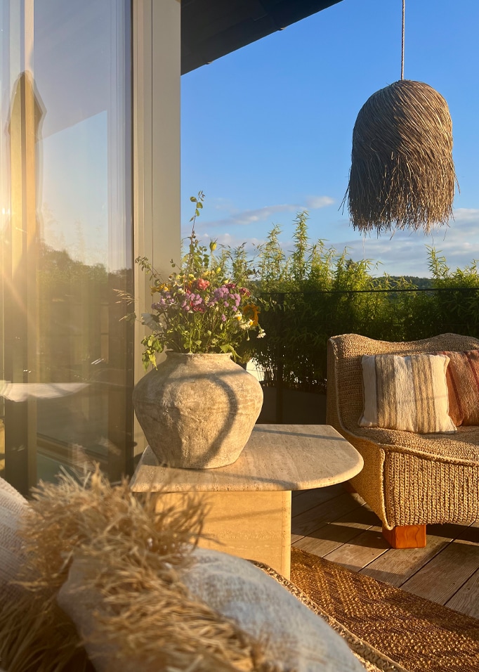 Przytulny balkon z wiklinowym fotelem, kamiennym stołem i wazonem z kwiatami w naturalnym świetle.